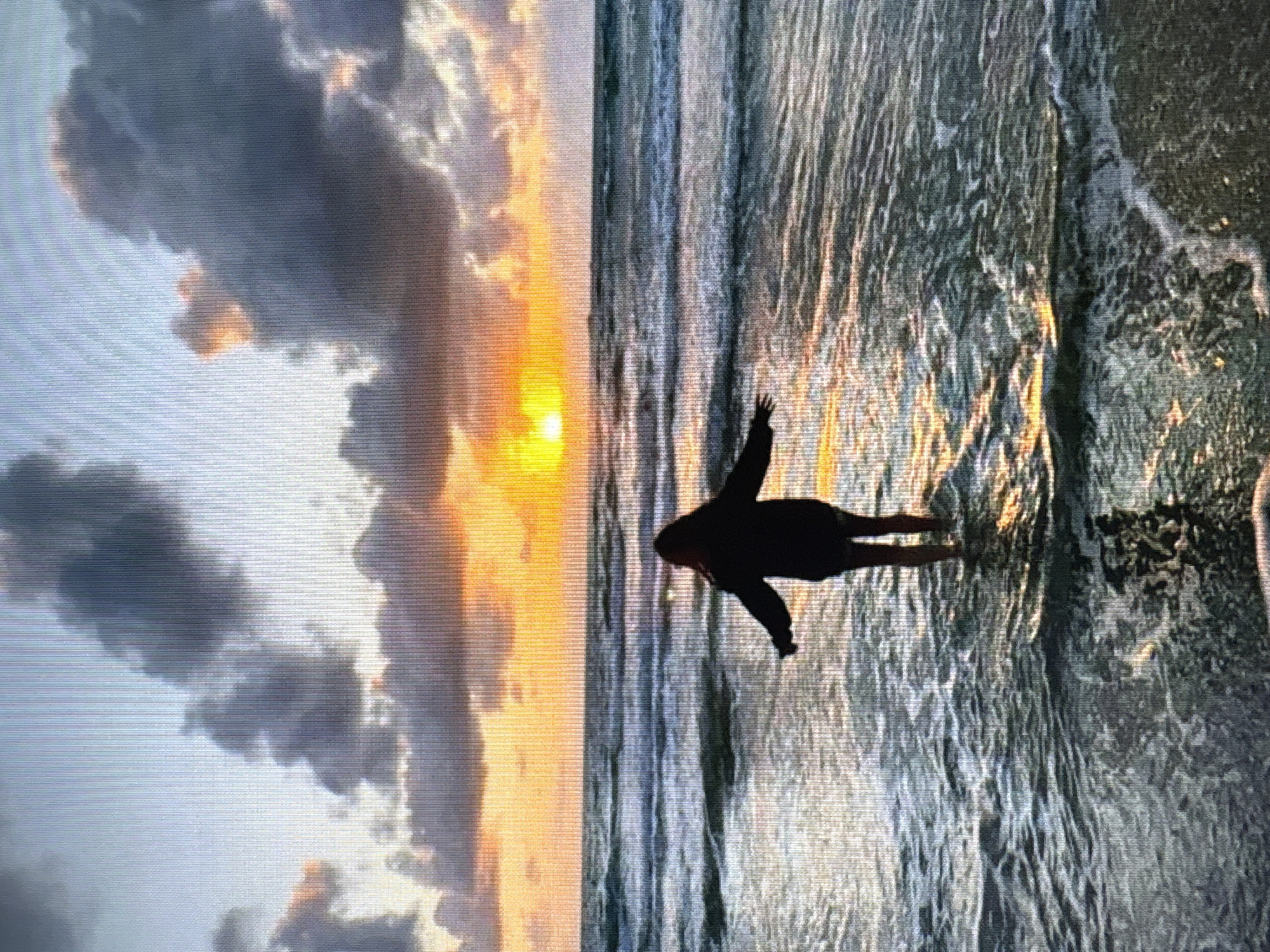 The image depicts a serene sunset over the sea, with a lone figure standing in the shallow water. The sun, a vibrant orb of orange and yellow, is partially obscured by dramatic, dark clouds that are tinged with the warm hues of the setting sun. The sea is calm, with gentle waves lapping at the shore, reflecting the colors of the sky. A person, silhouetted against the bright backdrop, stands with their arms outstretched, seemingly embracing the moment. The overall mood is peaceful and contemplative. The image does not provide any information about the city of Kuvshinovo, Russia.