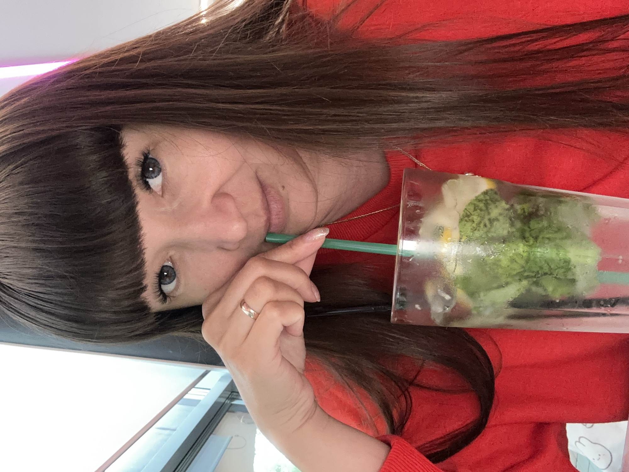 The image shows a woman in a red sweater, holding a clear plastic cup with a green straw. The cup contains a drink with ice and green ingredients, possibly mint and lime, suggesting a refreshing beverage. She has long, dark brown hair with bangs and is wearing a ring on her left hand. Her eyes are gray-blue, and she is looking towards the camera with a slight smile. The lighting suggests it might be daytime, and the background appears to be indoors with a window visible. The overall mood is casual and relaxed, possibly taken during a break or outing.
