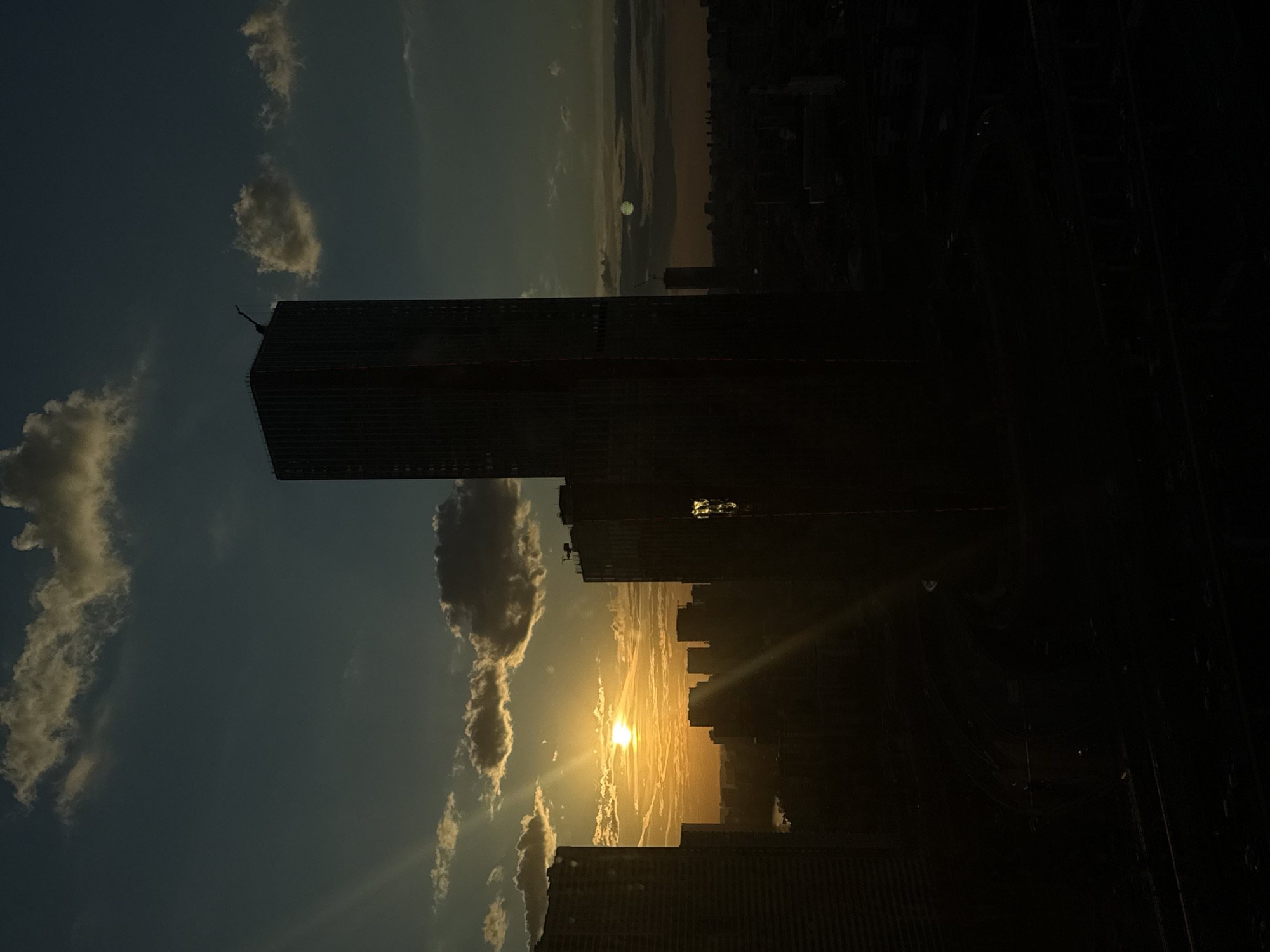 The media file displays an urban landscape at either sunrise or sunset, presenting a dramatic silhouette of buildings against a brightly lit sky. The image is taken from an elevated vantage point in what is understood to be Khimki, Russia.
The most prominent subjects are the numerous buildings, mostly appearing as dark silhouettes due to backlighting. A very tall, slender skyscraper dominates the left side of the frame, standing starkly against the vibrant sky. Other buildings form a dense, dark skyline extending across the mid-ground and into the distance. One window on a building to the right of the central skyscraper reflects a bright, yellowish light. The foreground is almost entirely black, obscuring any detailed structures or objects.
The central activity is the setting or rising of the sun, which is a brilliant yellow orb positioned low on the horizon, radiating intense light and creating a noticeable lens flare. The sky above the horizon transitions from bright orange and yellow directly around the sun to a deep, dark blue at the top. Scattered clouds, illuminated by the low sun, show warm hues of yellow and orange, adding texture to the sky.
There are no people visible in the image, and no discernible text. The overall mood is striking and atmospheric, emphasizing the interplay of light and shadow over a vast urban expanse.