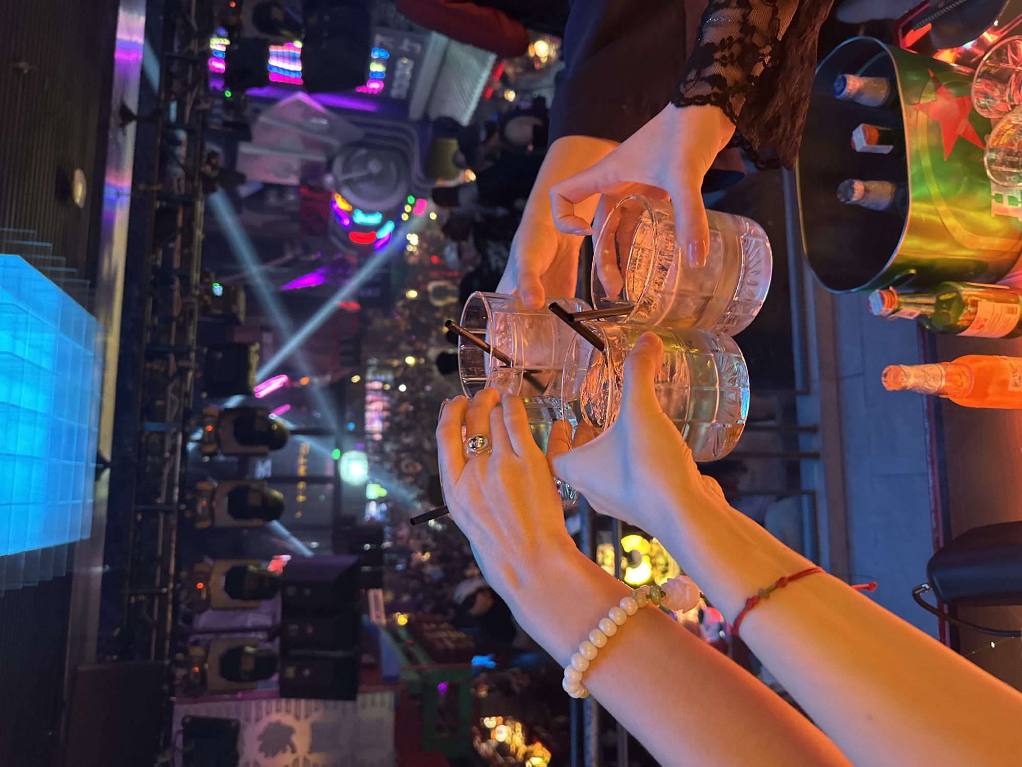 The image captures a group of individuals toasting with drinks in a dimly lit, vibrant nightclub setting. The scene is focused on their hands holding glasses filled with liquid and ice, with black straws. One hand wears a beaded bracelet and a ring. In the background, a crowded dance floor is visible, illuminated by colorful stage lights and moving beams of light, suggesting a lively atmosphere typical of a nightclub or bar. The overall mood is celebratory and social. There is no information to suggest the location is in Thủ Đức, Vietnam.