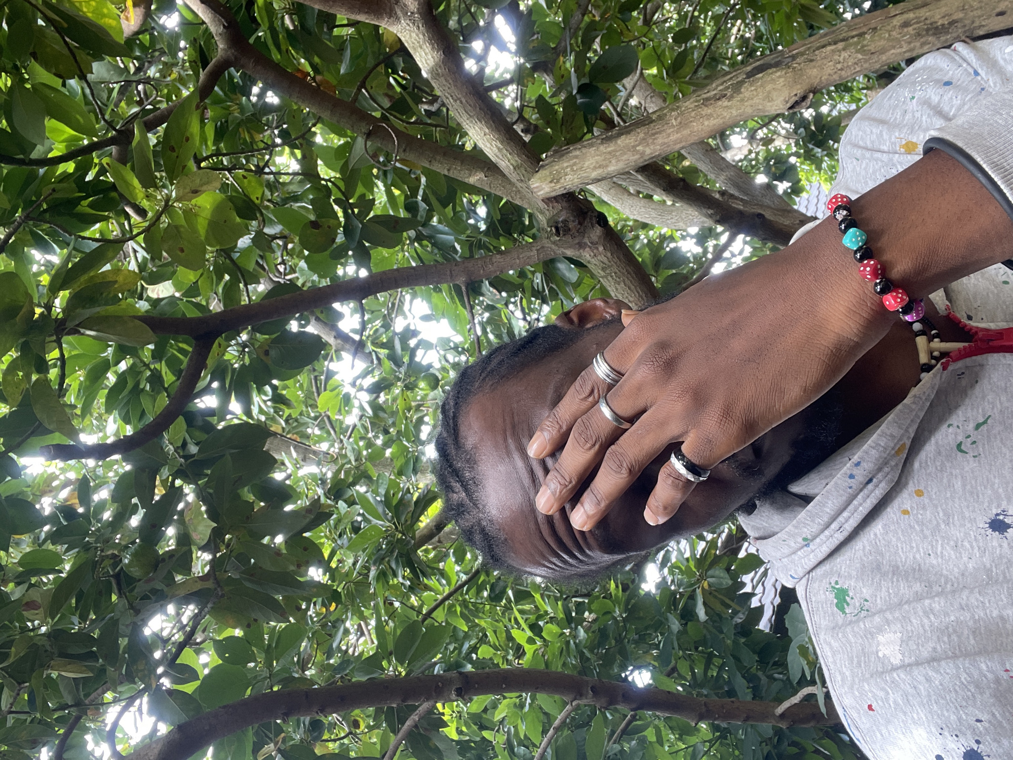 A medium shot shows a Black person covering their face with their right hand, amidst the branches of a tree. The person is wearing a grey hooded sweatshirt with multi-colored paint splatters and a bracelet on their left wrist made of colorful beads, some shaped like dice. The background is filled with green leaves and tree branches, suggesting an outdoor setting, possibly a garden or park. The person's skin is dark, and they have braided hair. The lighting suggests it is daytime, with sunlight filtering through the leaves. The specific location in Jos, Nigeria, cannot be determined from the visual information provided in the image.
