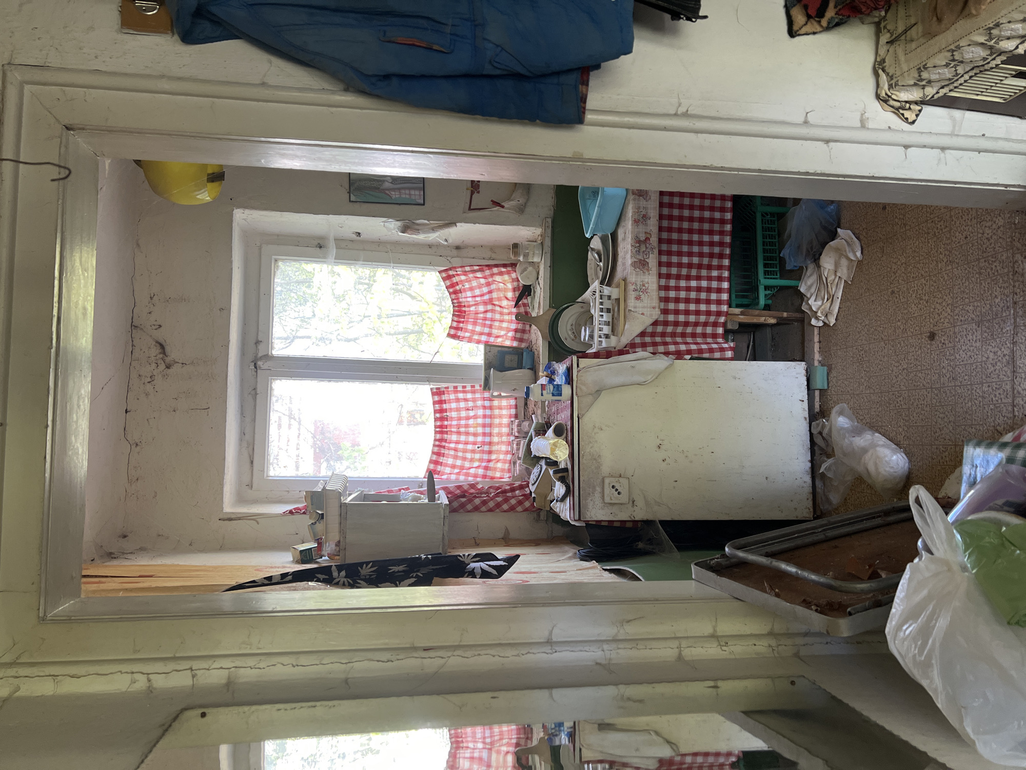 The image depicts a cluttered kitchen interior, viewed through a doorway. The kitchen features a window with red and white checkered curtains. There is a refrigerator, a table with a red and white checkered tablecloth, and various other items strewn throughout the space. The scene appears somewhat unkempt. The location is Boikovo, Bulgaria.