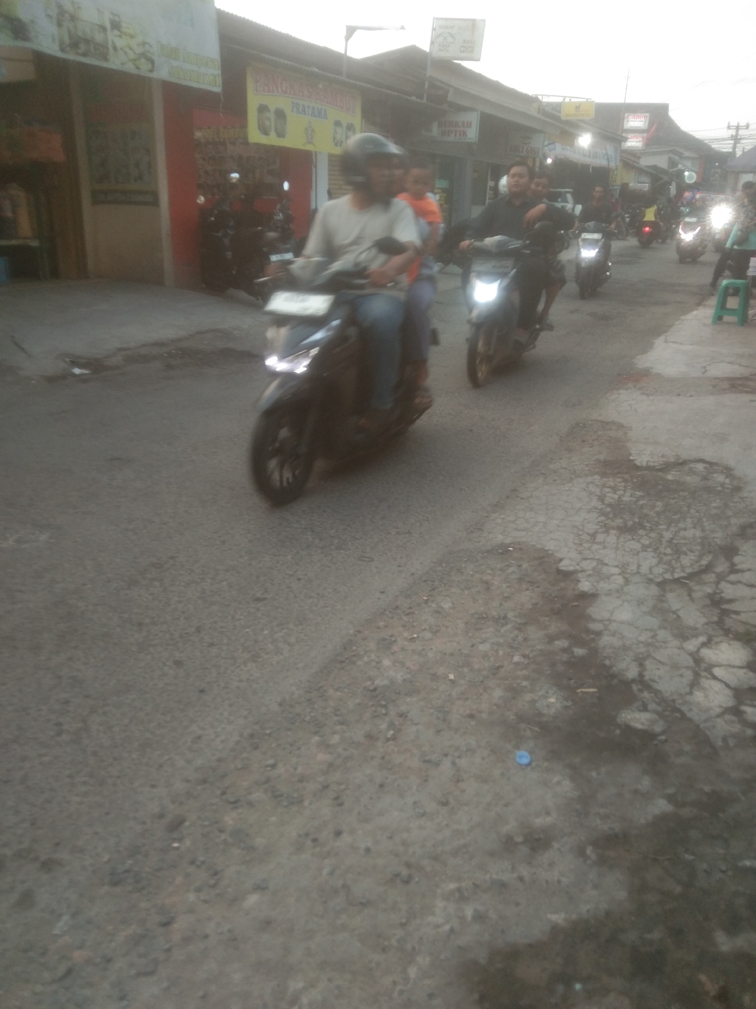 The image captures a street scene in Subang, Indonesia, likely during the day with overcast skies. Multiple motorcycles are present, with several individuals riding them. The road surface appears worn and cracked, with patches of what might be water or a different texture. Buildings line one side of the street, featuring signs that suggest commercial establishments, including one that reads "PANGKAS RAMBUT PRATAMA" (Pratama Hair Salon) and another possibly for optics. The overall atmosphere is that of a busy, everyday street in an Indonesian town.