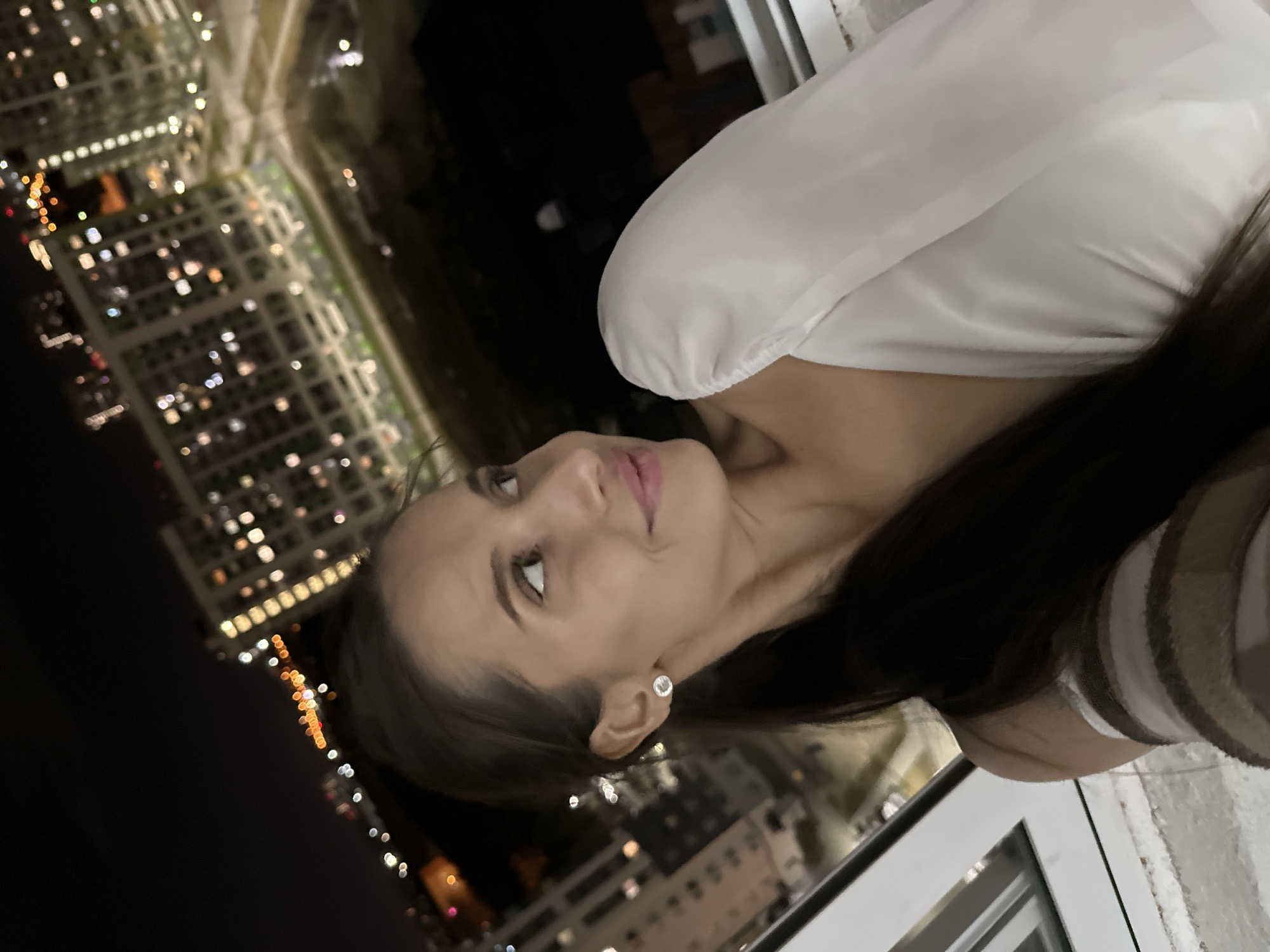 A woman is posed near a window in a high-rise building, likely at night. The scene overlooks a city with grid-patterned illuminated streets and buildings. The setting is possibly Yekaterinburg, Russia, given the urban environment. The woman is wearing a white blouse. She is looking upwards.