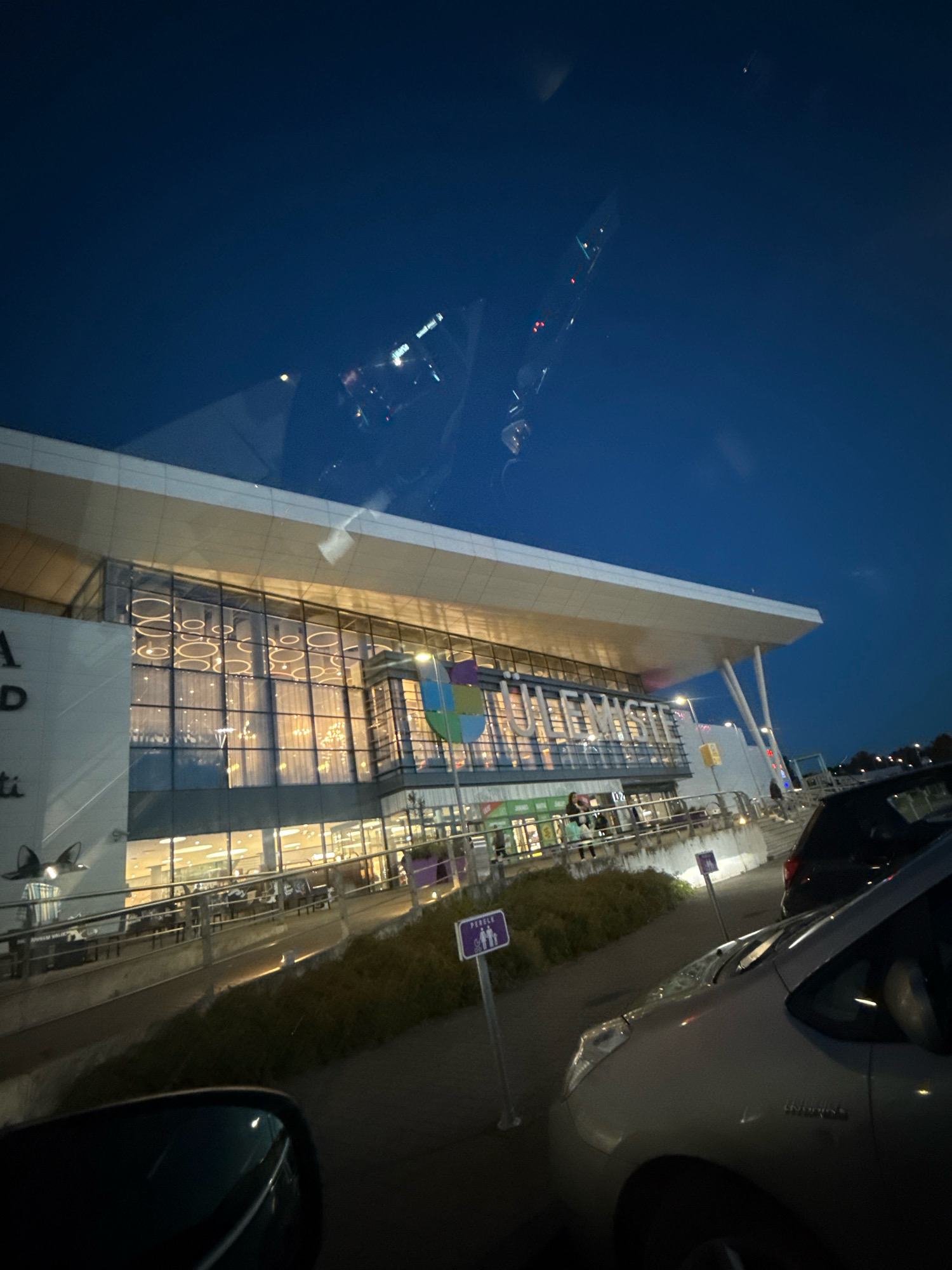 The image depicts the exterior of the Järve Keskus shopping center in Tallinn, Estonia, at dusk. The large, modern building features extensive glass facades illuminated from within, showcasing rows of circular light fixtures and curtains. A sign with colorful geometric shapes is mounted on the building. In the foreground, a silver car is partially visible, and the reflection of a car's interior can be seen in the side-view mirror. Two purple signs with pictograms indicating pedestrian crossings are positioned along the sidewalk. The sky is a deep blue, transitioning into darkness, with some indistinct reflections visible in the upper portion of the image.