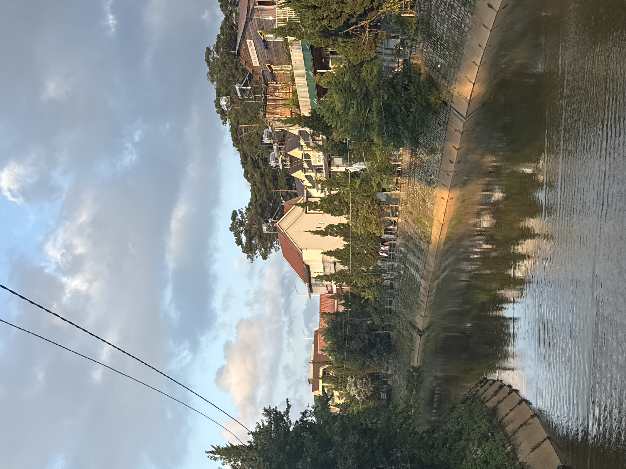 This image depicts a tranquil canal or small river flowing through a residential area within Phường Xuân Hương, Đà Lạt, Vietnam, under a partly cloudy sky. The lighting, characterized by warm, golden hues on certain clouds and buildings, suggests either early morning or late afternoon.
The upper portion of the frame shows a blue sky dotted with scattered grey and white clouds, some exhibiting a warm, sunlit glow. Two dark, parallel electrical or communication cables diagonally span the top-left quadrant of the sky.
The central and lower portions of the image are dominated by a narrow, calm waterway. The water is a dark, murky brown-green, reflecting the sky and surrounding structures, with visible ripples creating texture, particularly in the foreground. Both banks of the waterway are reinforced. The left bank is a sloping concrete embankment, partially overgrown with green vegetation and lined with dense, dark green coniferous trees.
The right bank is more developed, featuring a stone retaining wall topped with a railing. Behind this, a series of multi-story houses are constructed on a gentle incline, interspersed with numerous slender evergreen trees. The houses display diverse architectural styles, many featuring reddish-brown tiled roofs. Several houses have silver, cylindrical solar water heaters installed on their rooftops. A prominent wooden house on the far right exhibits a dark roof, a balcony, and a visible yellow sign with red Vietnamese text, which reads "Nhà Xe Dưỡng Thiện!". Further to the left, a building with a yellow facade displays a white sign with black text, "Ruby". Clothes are observed hanging on a line or railing adjacent to one of the houses on this bank, indicating human activity, though no individuals are visible. The hillside behind the houses is densely covered with pine trees, a characteristic feature of the Đà Lạt landscape.