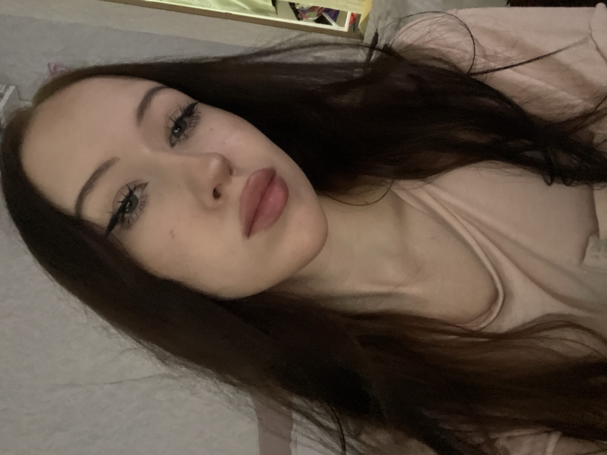 This is a close-up selfie of a young woman with long, dark brown hair, light skin, and dark eyeliner. She is wearing a light pink top. The background appears to be an indoor setting with a textured wall and a hint of a mirror or framed object to the right. There is no discernible indication of location within the city of Votkinsk, Russia, or any specific action or interaction.