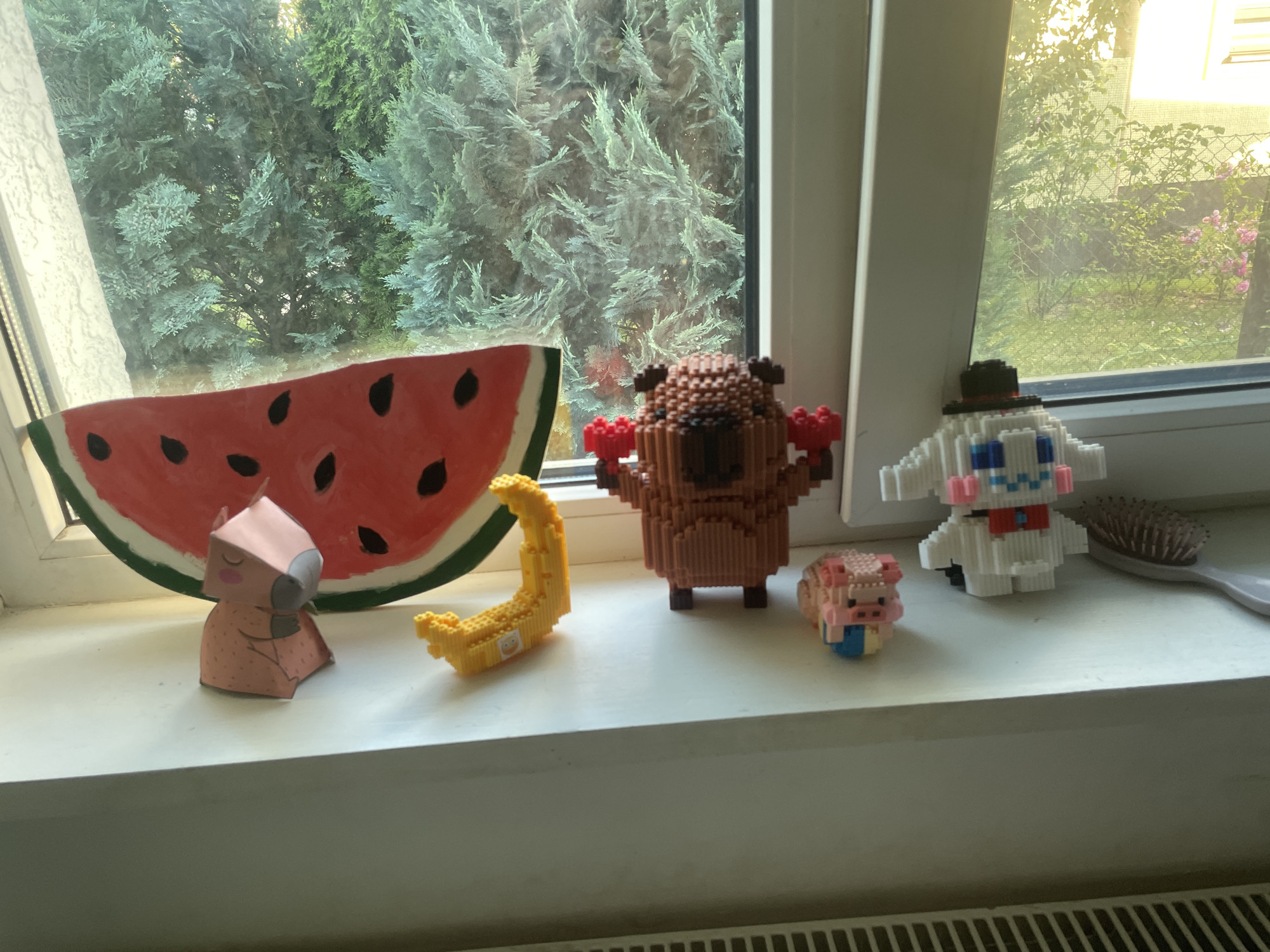 This media file features a collection of small, toy-like figurines arranged on a white windowsill. The figurines include a slice of watermelon, a yellow banana, a brown bear-like character holding red objects, a white character resembling Cinnamoroll from Sanrio wearing a black hat, a pink pig, and a pink papercraft animal. The windowsill overlooks a natural scene with lush green trees visible through the window, suggesting an outdoor setting. The lighting indicates it might be daytime, possibly morning or afternoon. The overall impression is one of a curated display of colorful objects. There is no visible text in the image.