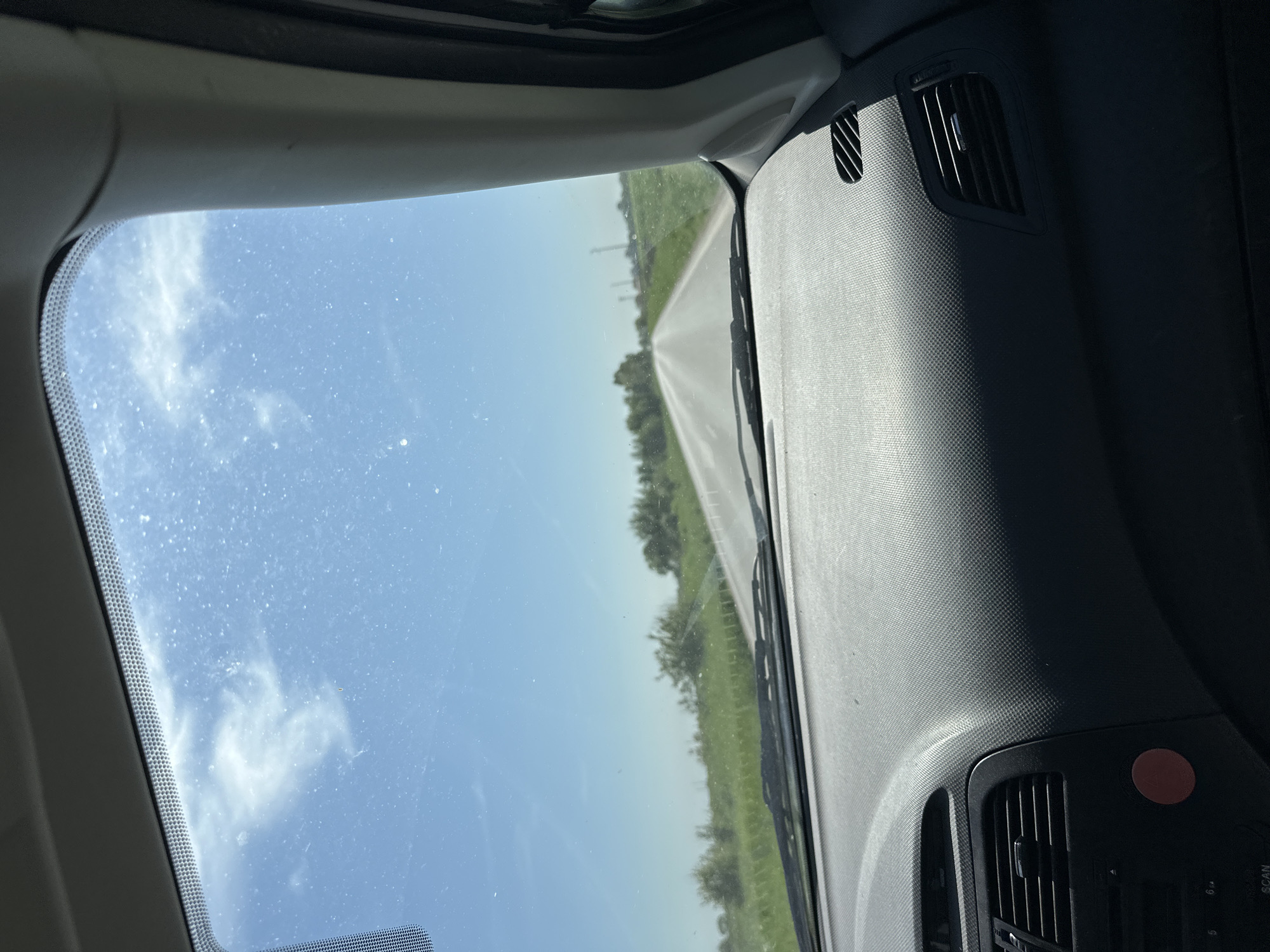 The image captures a first-person perspective from inside a car, looking out through the front windshield onto a straight rural road in Saedinenie, Bulgaria. The road extends into the distance, appearing to be a two-lane asphalt surface with a white center line. On either side of the road, there are expansive green fields and scattered trees, suggesting a countryside setting. In the distance, faint power lines can be seen against the horizon.
The sky above is bright blue with several scattered white, wispy clouds. The car's windshield is visible, showing numerous small dust particles or smudges across its surface. Inside the car, parts of the dark gray dashboard are visible on the right, featuring a textured material and air vents. Strong sunlight illuminates the dashboard, creating distinct highlights and shadows.
A portion of the car's A-pillar and headliner frames the view from the left side. At the bottom right of the visible dashboard area, a red circular button is present, along with a section of what appears to be an audio system control panel. Visible text on this panel includes "SCAN", "5", and "6". A segment of the windshield wiper blade is also seen at the base of the windshield on the right. The scene depicts a bright, sunny day, likely during a drive through the Bulgarian countryside. No people are visible.