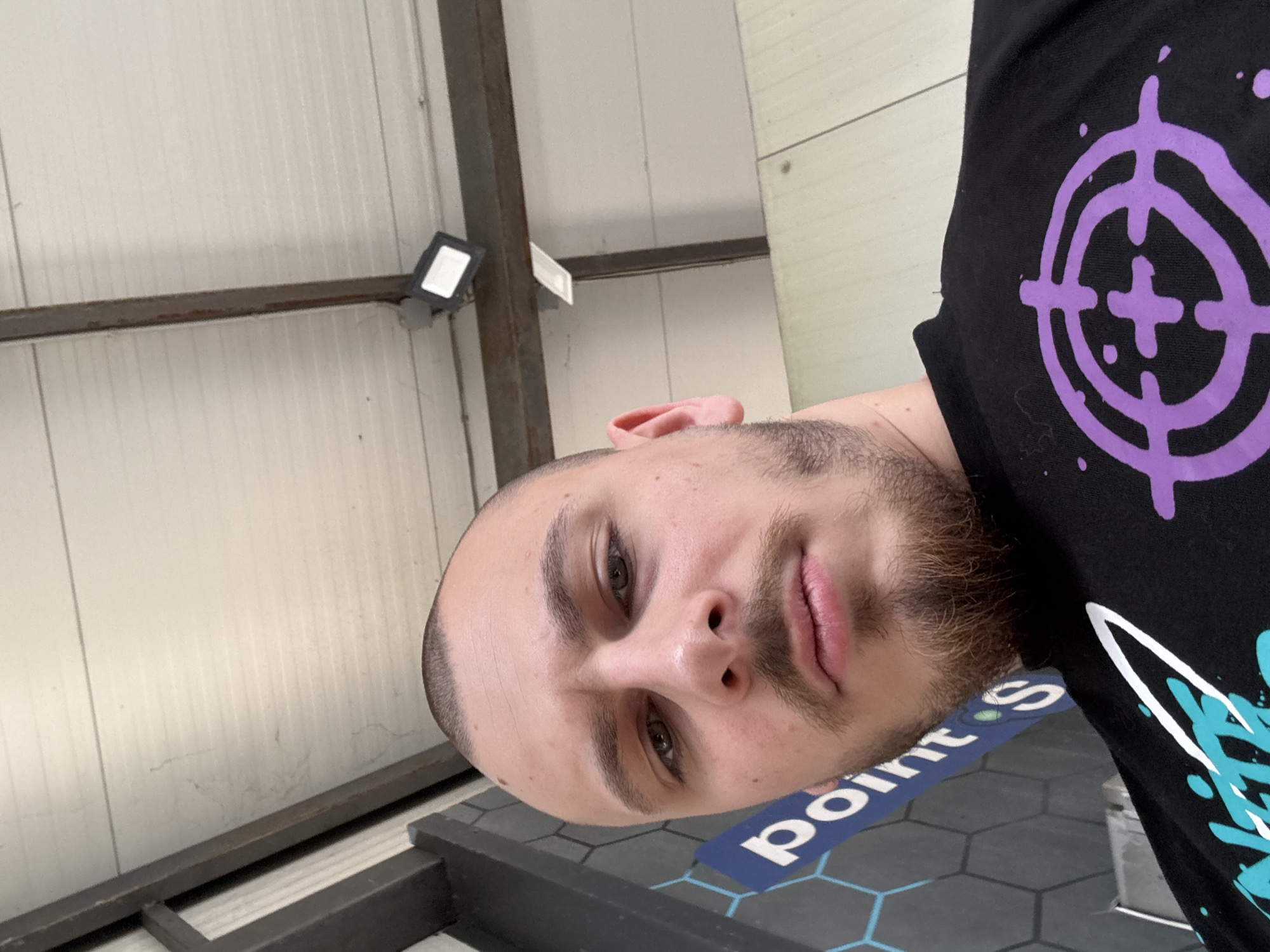 The image shows a man with short, light-brown hair, a beard, and hazel eyes. He is wearing a black t-shirt with a purple target symbol and a turquoise graphic design. He is standing on a surface with grey hexagonal patterns and the word "point S" printed on it. The background consists of corrugated metal sheets and a metal frame, possibly part of a building structure. A small rectangular light fixture is attached to the frame. The location might be an industrial or commercial space in the city of Stara Zagora, Bulgaria, given the "point S" flooring, which is a tire and automotive service company with locations in Bulgaria.