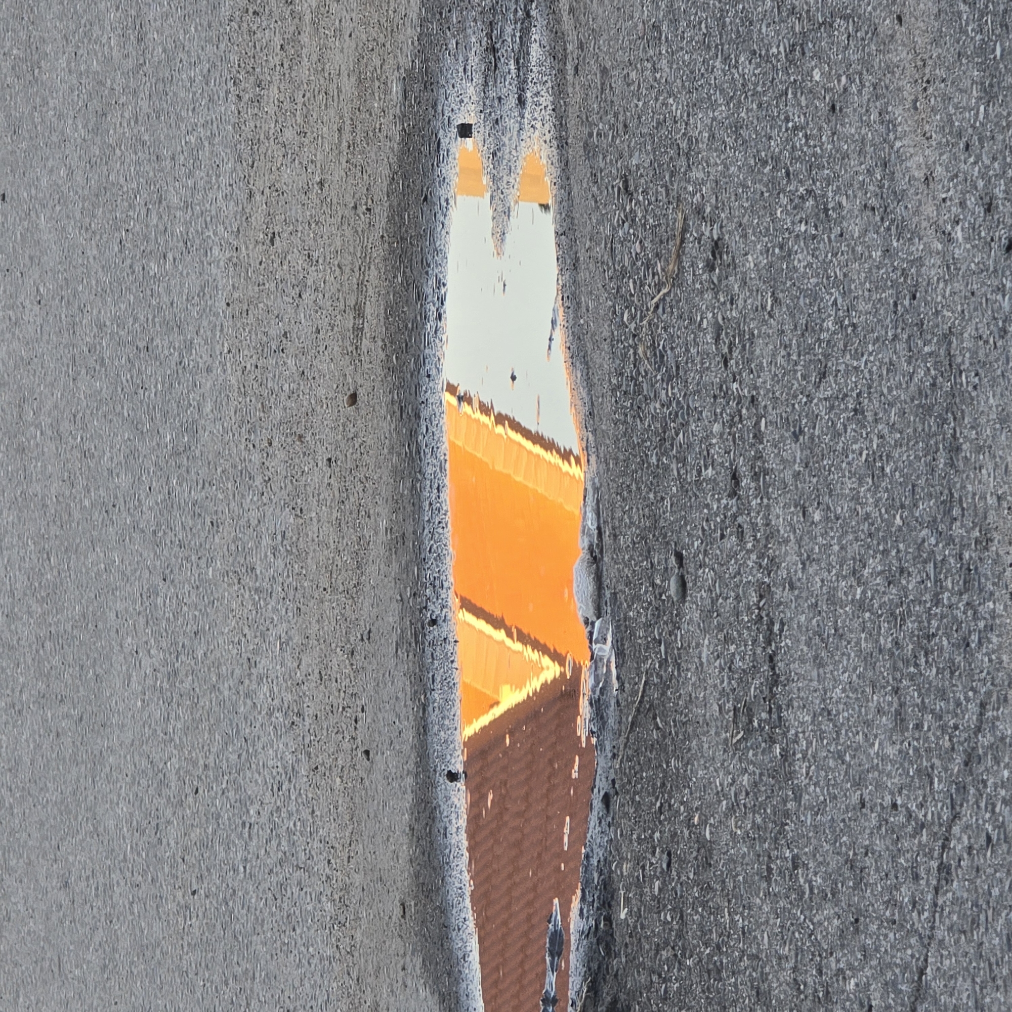A close-up, high-angle shot shows a puddle of water on a paved surface, likely a road or parking lot, in the city of Belokurikha, Russia. The puddle reflects the bright orange and brown colors of what appears to be a building with a distinct roofline, possibly a ski jump or a distinctive architectural feature. The surrounding pavement is gray and textured, with small pebbles and debris scattered across its surface. The water in the puddle is relatively still, with only slight ripples distorting the reflection. The lighting suggests it might be late afternoon or early morning, with the sun casting a warm glow on the reflected structure. There are no people or animals visible in the frame. The overall impression is one of urban decay and the beauty of an unexpected reflection.