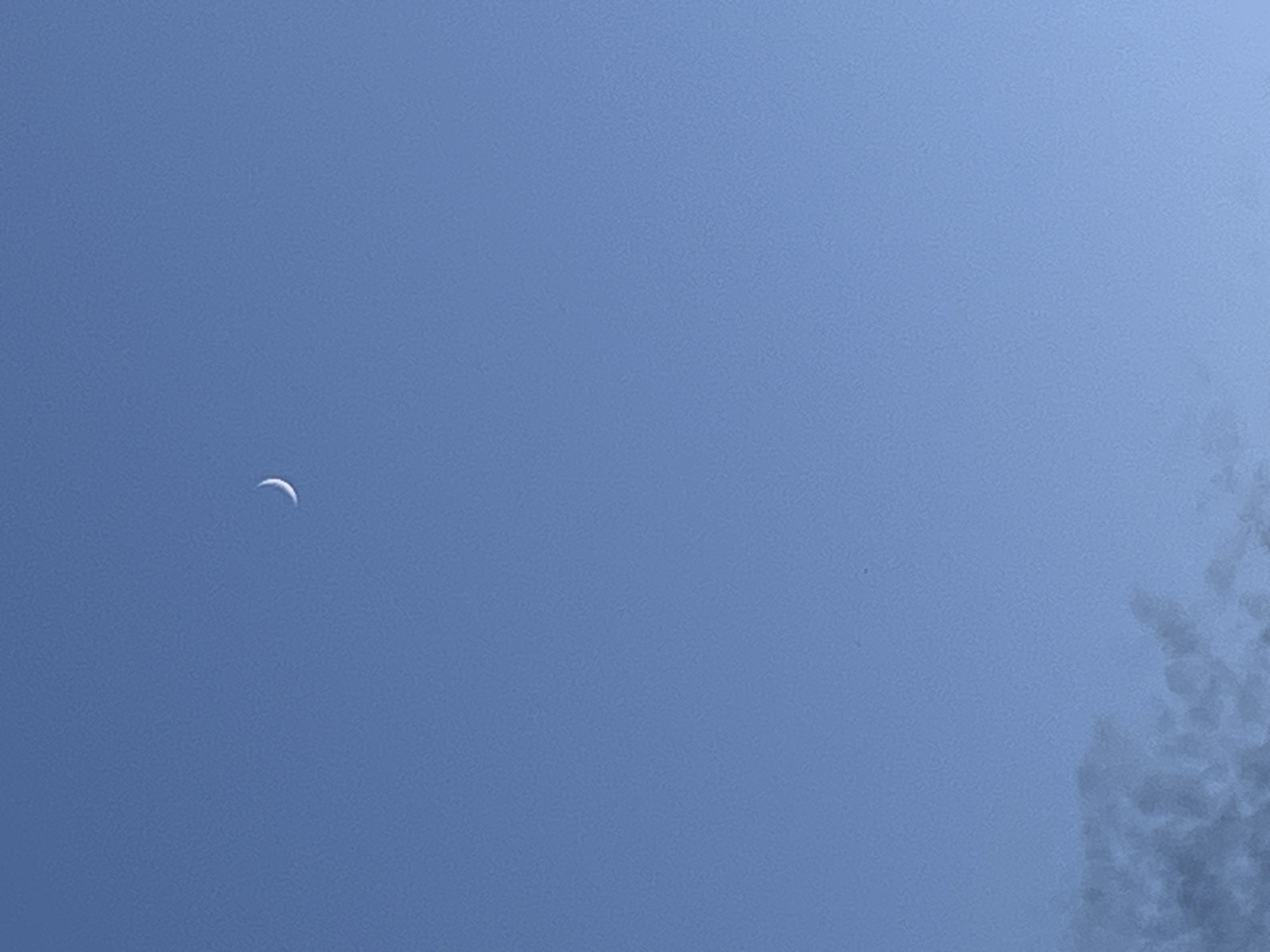 The image shows a crescent moon against a clear, blue sky. The moon is small and positioned in the upper left quadrant of the frame. To the right, the edge of what appears to be a cloud or smoke formation is visible, rendered in dark grey and blending into the sky. The overall scene depicts a daytime sky, with the moon visible due to daylight conditions and potentially its proximity to the sun. No people, objects, text, or specific activities are discernible. The setting is an outdoor sky.