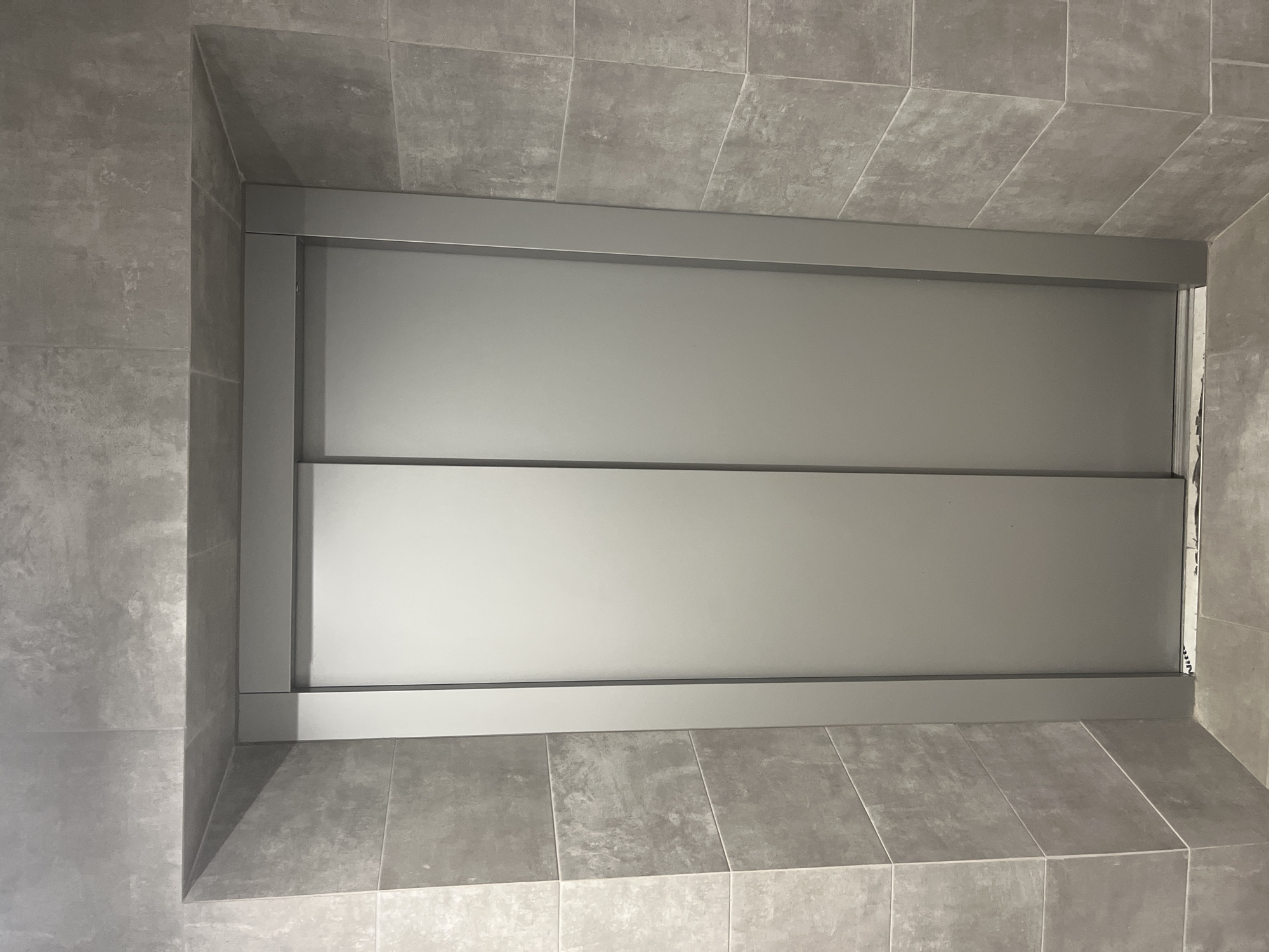 The image shows a gray-tiled alcove with a sliding door, likely a shower or storage area. The tiles are large and uniform in color, creating a seamless look. The sliding door has a silver frame. The location is unknown.