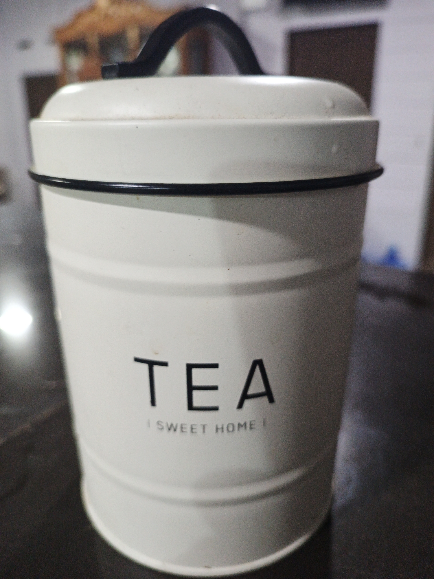 A white tin canister with a black handle sits on a dark, reflective surface. The word "TEA" is printed in black capital letters on the front of the canister, below which are the words "SWEET HOME" in smaller print. The canister has horizontal ridged lines and a black rim around its top. The background is blurred and shows indistinct shapes of furniture and walls, suggesting an indoor setting. There is no indication of the location being specifically in the city of Kelapapati, Indonesia.