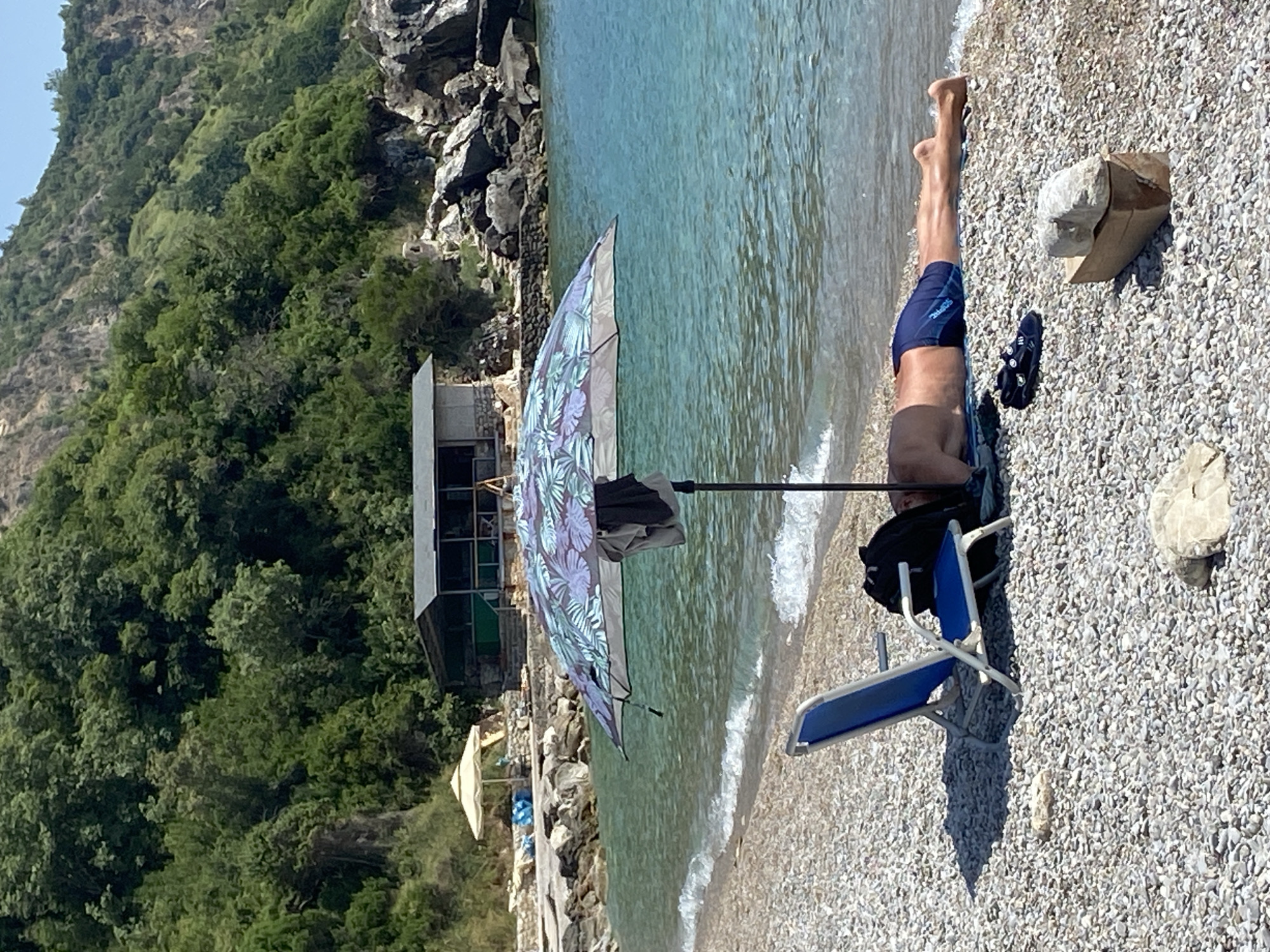A person is lying on a pebble beach in Prijevor, Montenegro, under a large, patterned umbrella. A blue beach chair is to the left of the person, and a black backpack is next to them, partially obscured by the umbrella. The person is wearing blue swim trunks and is positioned face down on a towel. A pair of black sneakers rests on the pebbles near their feet. The sea is clear and turquoise, with small waves lapping at the shore. In the background, there is a rustic building with a dark roof and stone walls, and beyond that, a densely forested hillside rises steeply. Rocks and vegetation are visible along the shoreline. The scene depicts a relaxed day at the beach under sunny conditions.