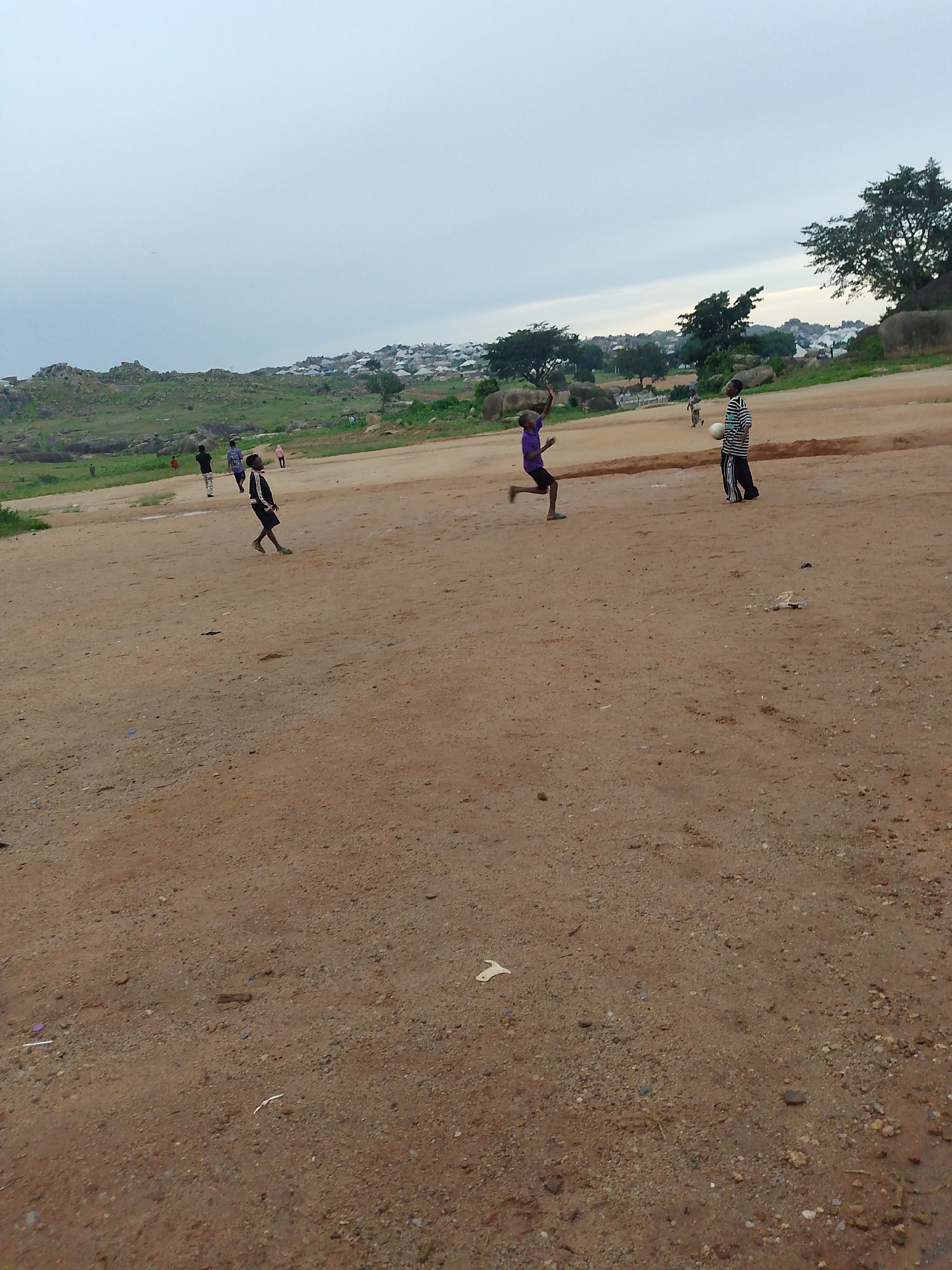 The image depicts an outdoor scene in Jos, Nigeria, featuring a dirt field with several young boys playing. In the foreground, a boy in a purple shirt is in motion, appearing to be kicking a ball. To his right, another boy in a black and white striped shirt stands holding a white ball. Further in the background, other individuals are visible, some walking and others playing, on the same dirt field. The field extends towards a slightly elevated area with sparse vegetation, rocks, and scattered buildings in the distance, suggesting a semi-urban or suburban environment. The sky is overcast, and the overall lighting indicates it is either early morning or late afternoon.