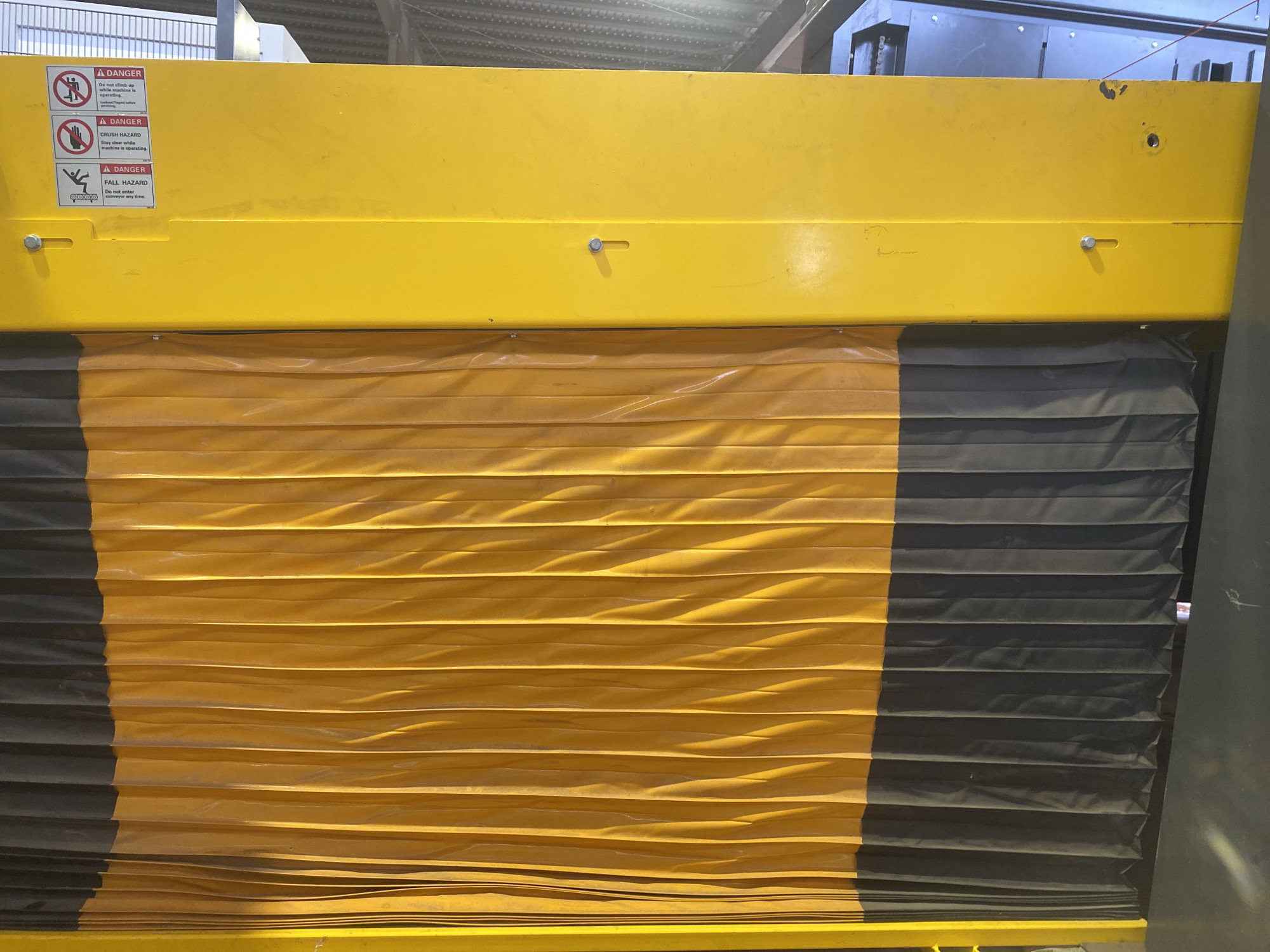 The image shows a piece of industrial equipment with a yellow and black striped curtain. The top part of the equipment is bright yellow and has three warning labels. From top to bottom, these labels are for "Do not climb up while machine is operating", "Crush Hazard", and "Fall Hazard". The curtain is made of alternating yellow and black horizontal stripes. The equipment is indoors, with the ceiling and some other equipment visible in the background. The location and the city of Paipe, Nigeria is not recognizable.