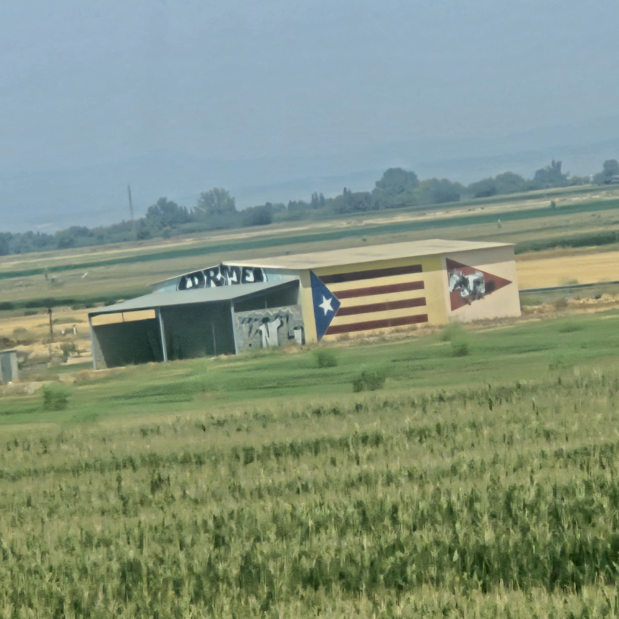 The image shows a long, rectangular building with a light-colored roof. The building is situated in a rural landscape, with fields of green and golden crops in the foreground and background. The building has two distinct sections: a darker, open-fronted structure on the left, and a more enclosed section on the right.
On the left section of the building, there is graffiti on the wall. The graffiti is in white and appears to be a stylized design with letters and figures.
The right section of the building features a mural. The mural consists of horizontal stripes in red and yellow, and a blue triangular section on the left side of the mural. Within the blue triangle, there is a white star. To the right of the stripes, there is another triangular section with a mural depicting what appears to be figures in a dynamic pose, possibly in motion or engaged in an activity. The overall colors of the building and its murals are somewhat muted, suggesting either a distance or a soft lighting condition.
The environment surrounding the building is agricultural, with rows of crops visible. In the distance, there are trees and hills under a hazy sky. A utility pole is visible to the left of the building, and a small, grey structure, possibly a shed or outbuilding, is located in front of the left section. The overall impression is of a functional building located in a farming area.