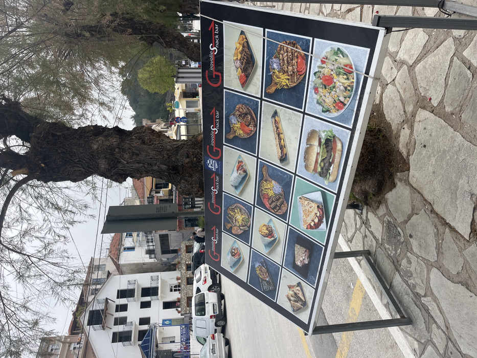 The scene shows a sign advertising a snack bar called "Girovollie Snack Bar", located in the town of Limenaria, Greece, at latitude 40.626 and longitude 24.577. The sign is mounted on a metal frame and displays a grid of 16 images showcasing various food items including gyros, burgers, and salads. The sign is positioned on a cobblestone pavement, partially shaded by a large tree trunk. The background shows several cars parked in a small, paved parking lot, with two people walking by. The buildings in the background are a mix of white, grey, and brown, with a few green trees and bushes visible. The sky is overcast.