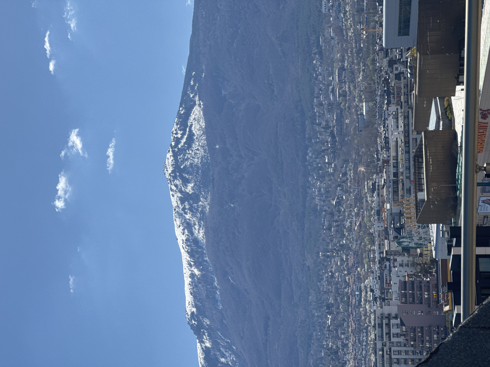The image depicts a cityscape with a large mountain range in the background, observed during daytime with a clear blue sky and some scattered clouds. The mountain range is partially covered with snow. The cityscape consists of various buildings of different heights and architectural styles.
It is strongly suggested that this photo was taken in Sofia, Bulgaria, with Vitosha Mountain in the background.