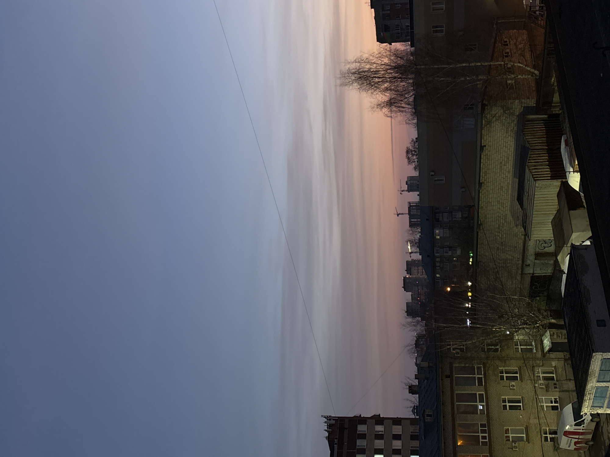 The image shows a dusk or dawn cityscape in Perm, Russia, taken from an elevated position, likely a balcony or high-rise window. The sky is gradated, transitioning from blue in the lower left to orange near the horizon line. Tall apartment buildings are visible, interspersed with trees. Utility lines stretch across the sky. Buildings are lit by streetlights and internal building lighting. The lower right shows a close-up view of the building with air-conditioning units on the facade and a partially visible white van parked below. Construction cranes appear in the distance.