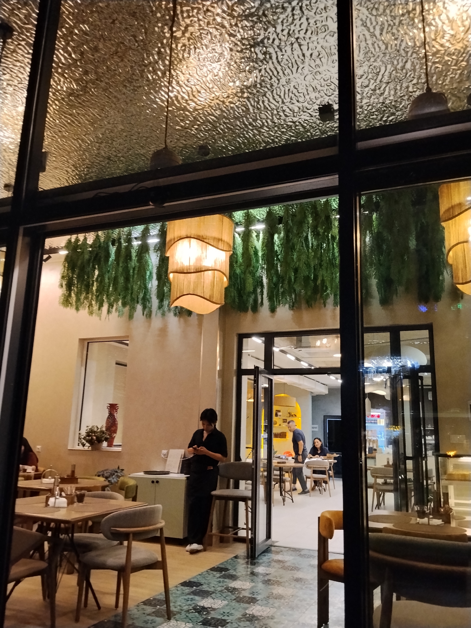 This media file depicts the interior of a modern, dimly lit restaurant or cafe in Bishkek, Kyrgyzstan. Through a large glass facade, we see several tables and chairs arranged for dining. Hanging plants add a touch of greenery to the ceiling, and unique, layered lamps provide warm illumination.
Several people are visible within the establishment. In the foreground, a person wearing a dark uniform, likely a server, is standing and looking at their phone. Further inside, a man and a woman are seated at a table, appearing to be engaged in conversation or work. Other patrons are dimly visible at tables in the background.
The overall ambiance suggests it is evening. The textured glass ceiling reflects the interior lights, creating an interesting visual effect. The flooring features a decorative tiled pattern. A large vase with a red, ornate design is placed on a shelf near a window, adding to the restaurant's decor. The scene captures a typical evening at a contemporary dining establishment.