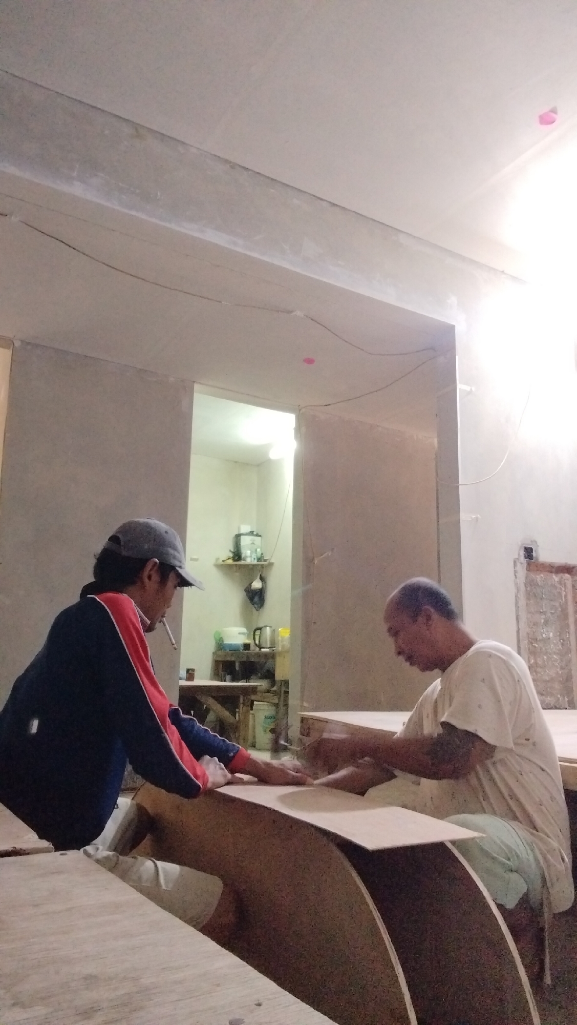 Two men are working on unfinished plywood furniture in an indoor workshop. The man on the left, wearing a grey baseball cap, blue and red jacket, and white shorts, is smoking a cigarette and placing his hands on a curved piece of plywood. The man on the right, wearing a light-colored t-shirt and light blue shorts, is also working with plywood. The background shows an open doorway leading to a small, sparsely furnished room with shelves and a kettle. The walls of the workshop appear to be unfinished, with exposed beams and electrical wiring visible on the ceiling. The lighting is dim, with a bright light source coming from the doorway. The overall scene suggests a construction or renovation site.