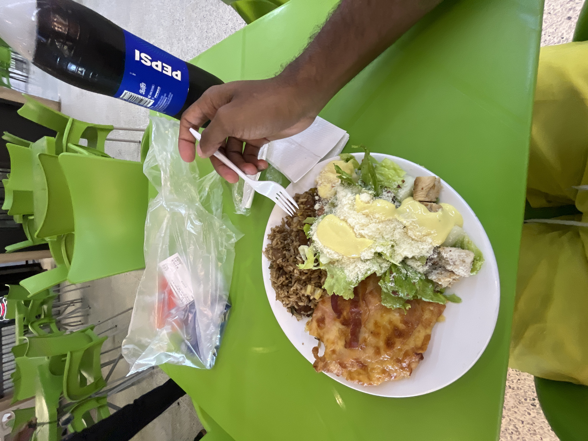 The image captures an overhead view of a person about to eat a meal at a bright green table in what appears to be a cafeteria or food court setting in Porlamar, Venezuela. A dark-skinned arm and hand, holding a white plastic fork, are positioned over a white plate of food.
The plate holds a diverse meal: a substantial portion of Caesar salad with fresh green lettuce, croutons, a creamy yellow dressing, and ample grated white cheese. Alongside the salad, there is a serving of brownish rice mixed with beans or lentils. Completing the meal is a piece of meat, possibly chicken, topped with a golden-brown melted cheese or sauce.
A large, dark plastic bottle of Pepsi is visible in the upper left corner, partially filled with the beverage. The blue label on the bottle clearly shows the brand name "PEPSI" in white capital letters. Underneath the person's hand, a stack of white paper napkins rests on the table. To the left of the plate, a clear plastic bag contains small, indistinct items, possibly condiments or extra cutlery.
The table and surrounding chairs are a vibrant lime green, with some chairs stacked, indicating a public dining area. The floor in the background is a light, speckled grey. The scene depicts a casual mealtime. No specific time of day or emotional state is discernible.