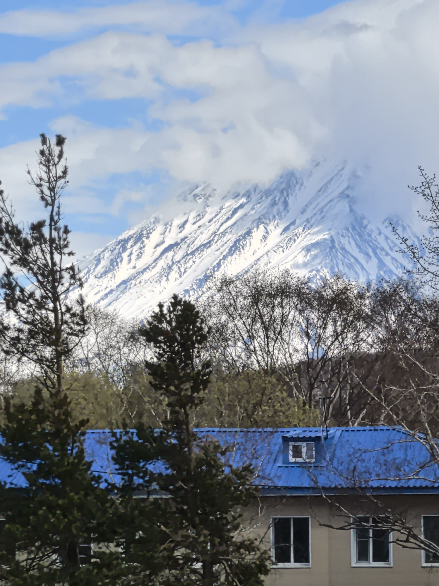 The image captures a snow-capped mountain obscured by clouds, set against a backdrop of a blue sky with scattered clouds. In the foreground, a building with a blue roof and several windows is partially obscured by trees with bare branches. The scene is likely located in the city of Unknown, Unknown, given its description in the prompt. The landscape exhibits a contrast between the natural environment of the mountain and trees, and the man-made structure of the building.
