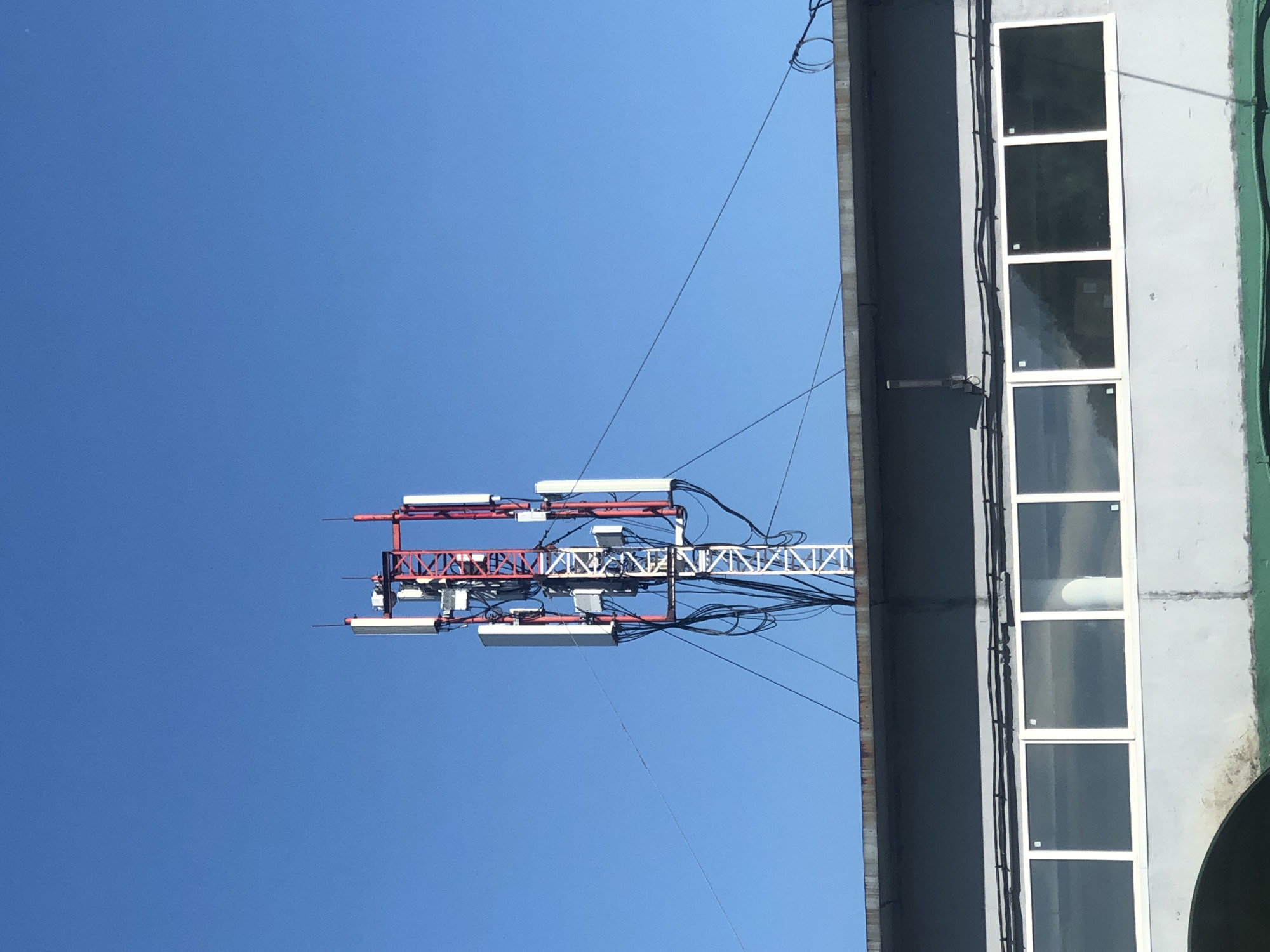 The image shows a cell tower with multiple antennas against a clear blue sky. The tower is situated behind a grey building with a row of large, rectangular windows. The building's roofline is visible in the foreground, and power lines extend from the tower to the building and other unseen structures. The scene appears to be an urban or industrial area.