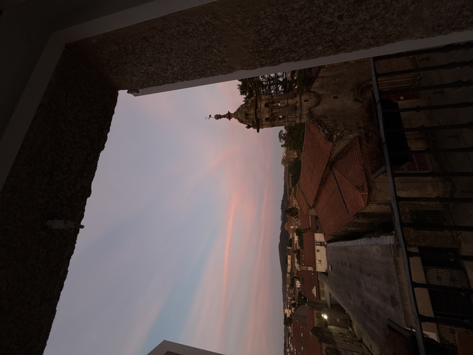 The image shows a view of Santiago de Compostela, Spain, during sunset. The sky is painted with soft hues of pink and blue. In the foreground, a balcony railing with dark metal bars partially obstructs the view. Beyond the railing, a cluster of buildings with terracotta tile roofs are visible. To the right, a stone bell tower with a spire, adorned with a cross, stands prominently. The lower section of the image reveals the dark rooftops of houses and what appears to be a section of a church. The environment suggests a historical urban setting. There are no people visible in the image. The lighting indicates it is dusk.