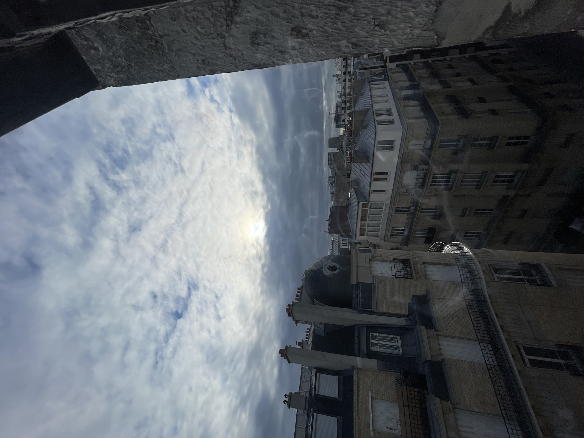 The image depicts a view of Parisian rooftops under a cloudy sky with the sun partially obscured. The architecture suggests a historic district in Paris, characterized by mansard roofs, dormer windows, and ornate balconies, typical of Haussmannian buildings. The foreground features a rough-hewn stone structure, likely part of a monument or older building, framing the cityscape on the right. In the middle ground, several buildings are visible, some with dark grey roofs and chimneys, others with lighter facades and windows, some featuring closed shutters. The sky is a mix of grey and white clouds, with a diffused sunlight breaking through, casting a soft light on the scene. There are no people or discernible actions taking place. The overall impression is one of an overcast day in an urban environment.