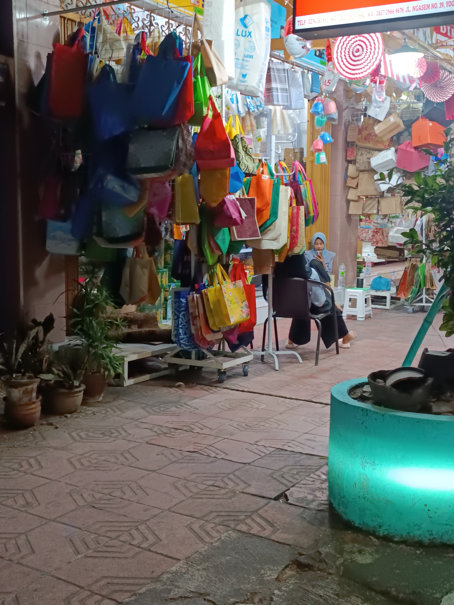 The image depicts an outdoor stall in Yogyakarta, Indonesia, likely a marketplace or a roadside vendor. The stall displays a wide assortment of colorful reusable shopping bags, hanging from a metal rack. Some bags are solid colors like red, blue, and green, while others have various patterns. In the background, more bags and decorative items like paper fans are visible, along with some cardboard boxes and a sign that appears to be a price list or advertisement.
A woman wearing a blue hijab and dark clothing is seated on a dark chair, looking at her phone. She is positioned in front of the stall, with a light-colored stool and a water bottle nearby. Another person is partially visible, seated in front of her.
The ground is paved with interlocking reddish-brown tiles with a geometric pattern. In the foreground, there is a large, circular, turquoise planter with a bright green light emanating from it, illuminating the lower right portion of the image. Several potted plants are arranged on a low platform to the left of the stall, adding a touch of greenery to the scene. The overall impression is of a busy commercial area, likely at dusk or in the evening, given the lighting.