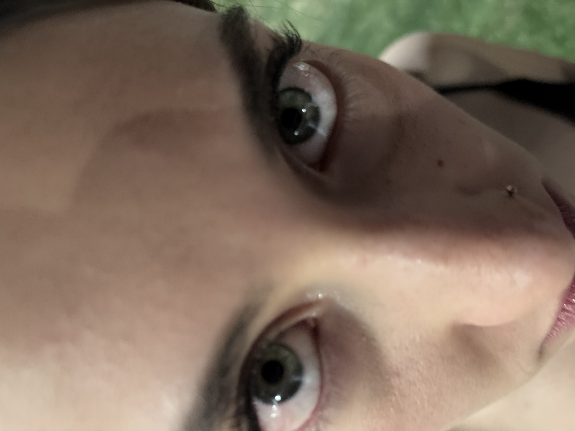 Close-up of a person's face, angled upwards. The subject has fair skin, green eyes, and a nose piercing. Their eyebrows are dark. A portion of their lips is visible. The setting is unclear but appears to be outdoors with a blurred green background. No specific location within Huixquilucan de Degollado, Mexico, can be determined from the image.