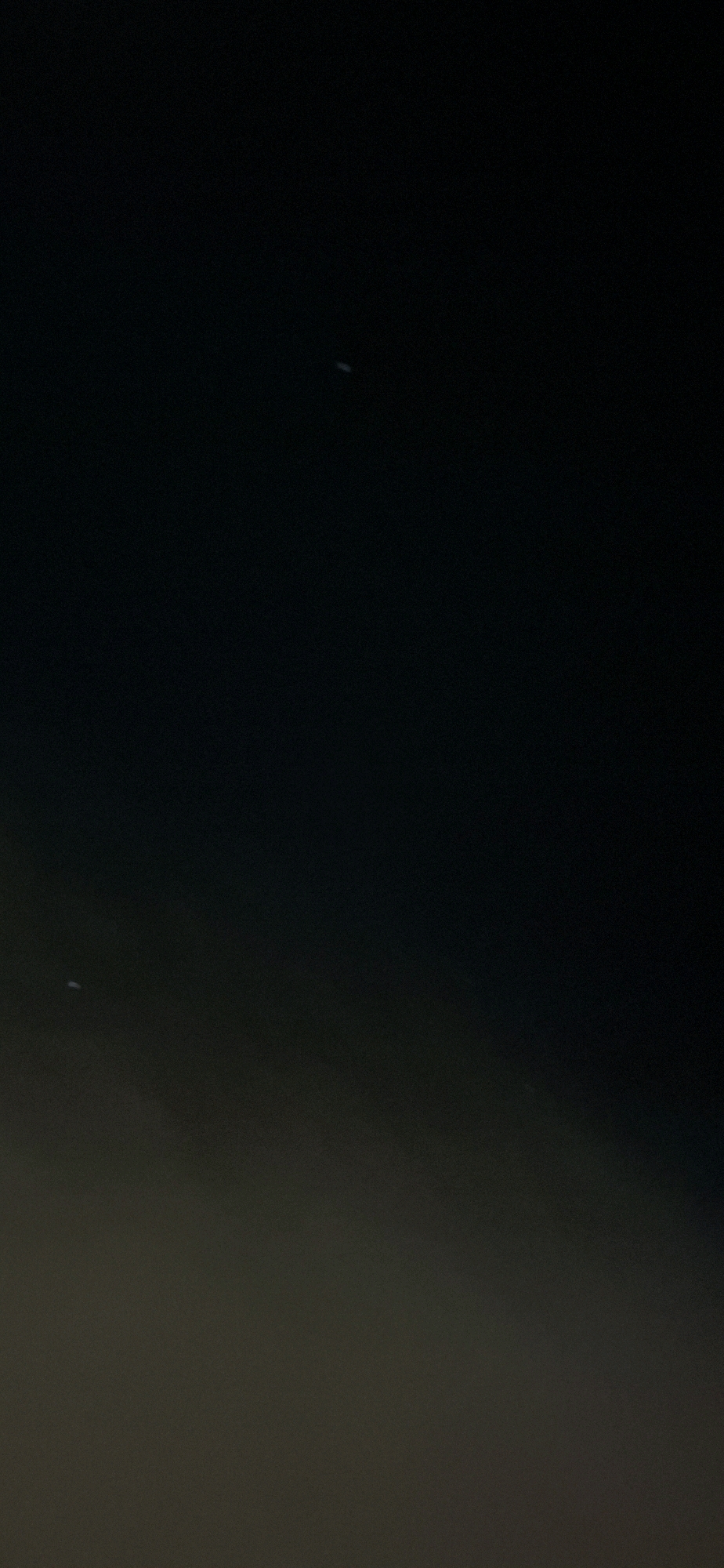 The image shows a night sky with two small, white, blurry objects that appear to be lights or stars. One object is located in the upper right quadrant of the image, and the other is in the lower left quadrant. The sky is dark, with a gradient from black at the top to a lighter, grayish hue at the bottom, suggesting some atmospheric haze or light pollution. The scene is devoid of any discernible landmarks or context that would indicate its specific location within Yerocham, Israel.