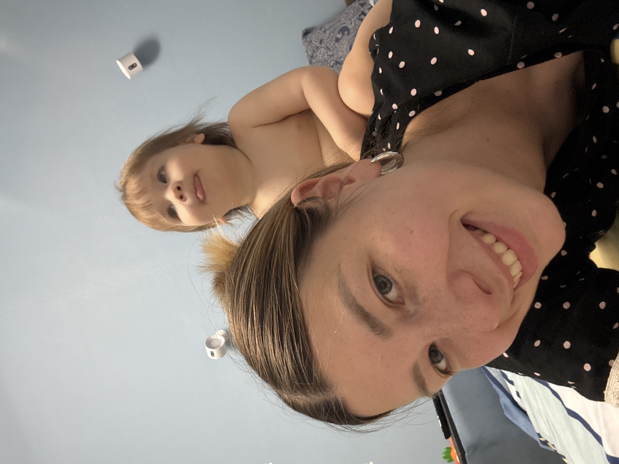 A woman and a child are pictured indoors. The woman is smiling at the camera while holding the child in her arms. The child is looking towards the camera with their tongue sticking out. The woman is wearing a black top with white polka dots. There is a white object mounted to the wall in the background. The location is not discernible from the image.