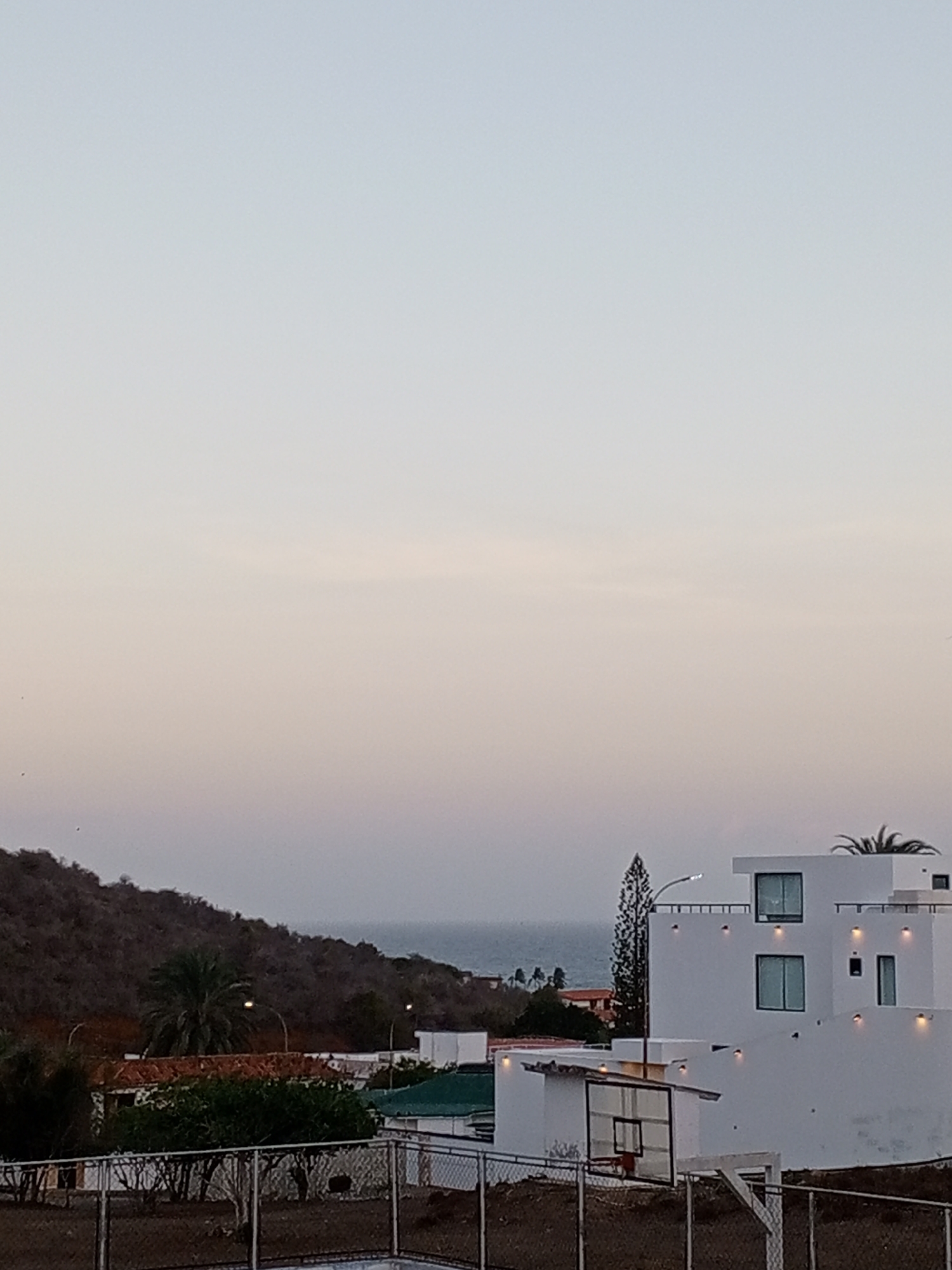 The image depicts a view in Pampatar, Venezuela, at dusk or dawn, characterized by a pale sky. A hill covered in vegetation is visible in the background on the left. The sea is faintly visible behind the hill. In the foreground, there's a white, multi-story building with a modern architectural style, featuring square windows and a flat roof. A basketball hoop is situated on a lower level of the property, partially obscured by a fence. Vegetation is scattered around the scene. There are no people in the scene. The focus is on the architecture and landscape of the area.