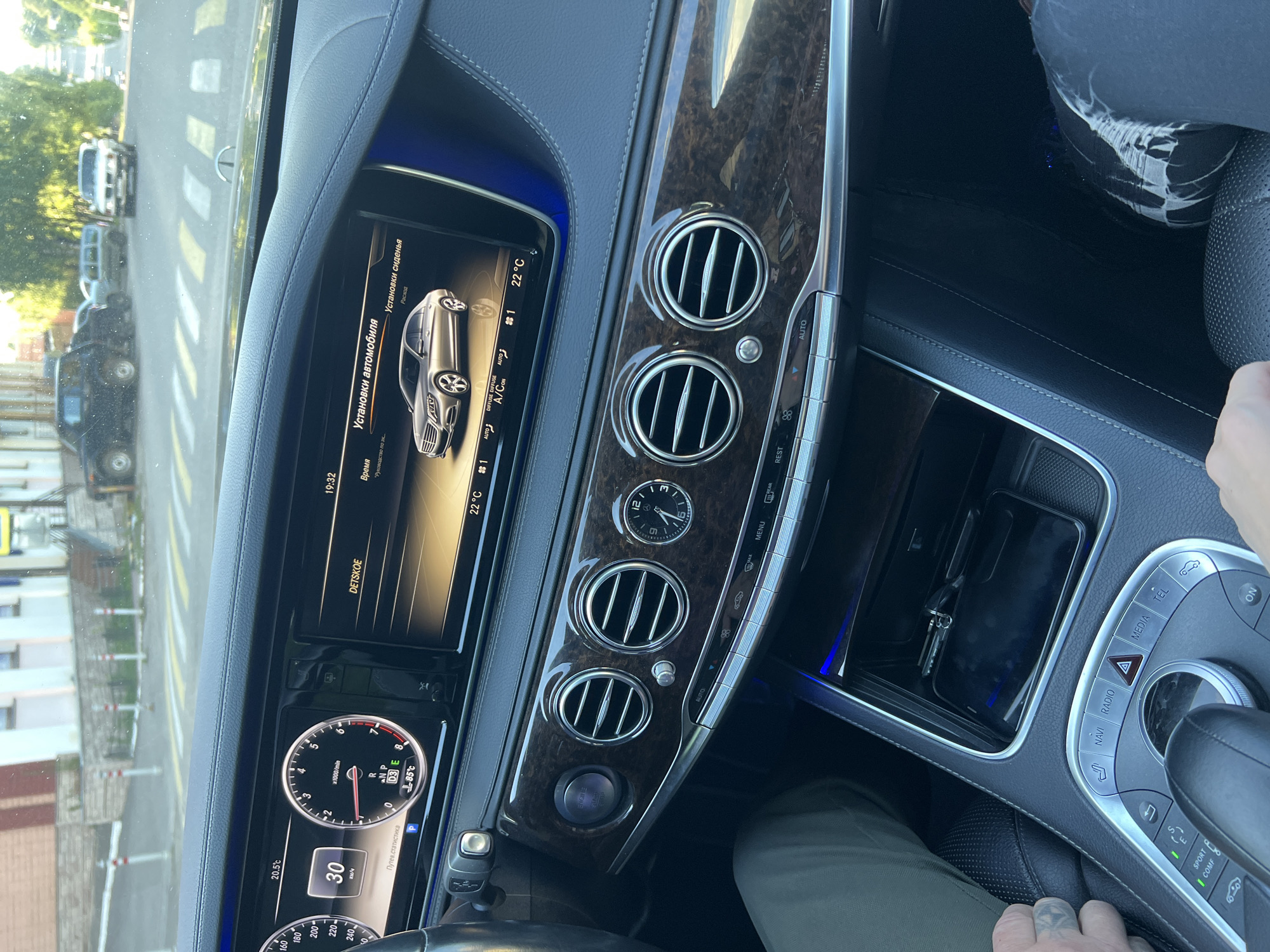 The image shows the interior of a luxury car, likely a Mercedes-Benz S-Class, with a focus on the dashboard and infotainment system. The car is parked on a street in Krasnoe-na-Volge, Russia, as indicated by the visible street signs and architecture.
In the foreground, a hand with tattoos is visible on the left, resting on the car's perforated leather seat. On the right, a person's legs, clad in dark pants with a distressed pattern on the right leg, are visible.
The dashboard features a prominent digital display showing vehicle information, including speed (30 km/h), engine RPM, temperature (20.5°C and 22°C), and various system settings such as "ДЕТСКОЕ" (Children) and "УСТАНОВКИ АВТОМОБИЛЯ" (Vehicle Settings). A silver sedan is depicted on the screen, along with climate control information.
The center console has wood trim, circular air vents with chrome accents, and several buttons and controls. The start/stop engine button is visible and illuminated. Below the console, a phone and keys are placed in a storage compartment.
The exterior view through the windshield reveals a street scene with buildings, trees, and other vehicles, including SUVs and a pickup truck, typical of a small Russian town. The lighting suggests it is daytime, possibly late afternoon, with the sun casting shadows. The environment appears to be a public road in a residential or commercial area.