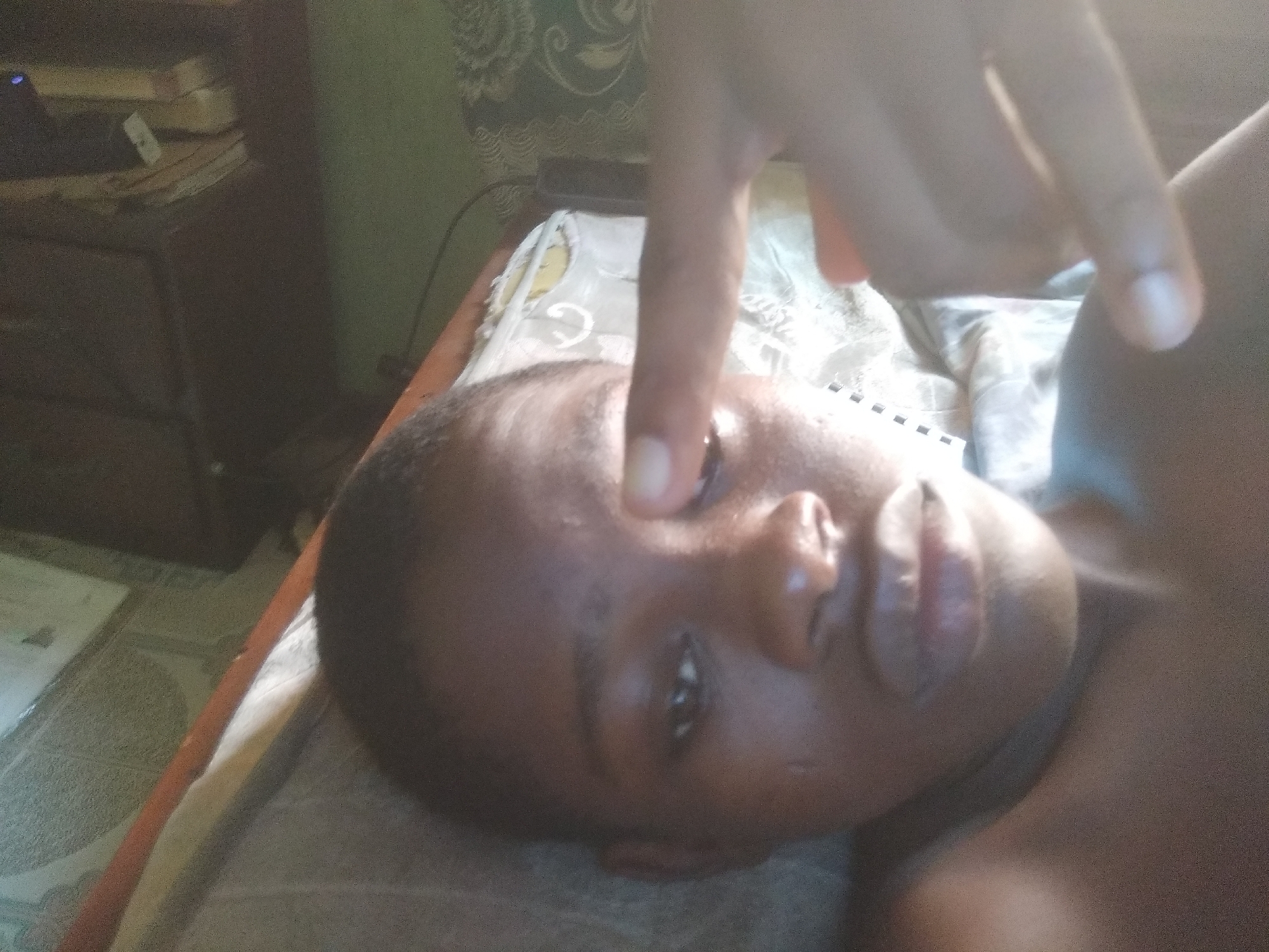 An eye-level shot captures a person with dark skin tone lying on their back, seemingly on a bed or cushioned surface. Their head is tilted to the right, presenting their face prominently in the foreground. An index finger from an unseen hand extends downwards, pointing directly towards the person's left eye, almost touching it. The person's left eye is open, and there is a visible sheen or moisture around it. Their expression appears neutral, or slightly uncomfortable due to the finger's proximity to the eye.
The background reveals an indoor setting. To the left, a dark wooden piece of furniture, possibly a bedside table or chest of drawers, is visible with what looks like stacked books or papers on top, and a small device emitting a faint blue light. The wall behind has a subtle, light-colored patterned wallpaper. The person is dressed in what appears to be a dark or sleeveless top that blends with their skin tone. The lighting is diffused, suggesting an indoor environment, possibly daylight coming from a window or general room lighting. No text is visible in the image.