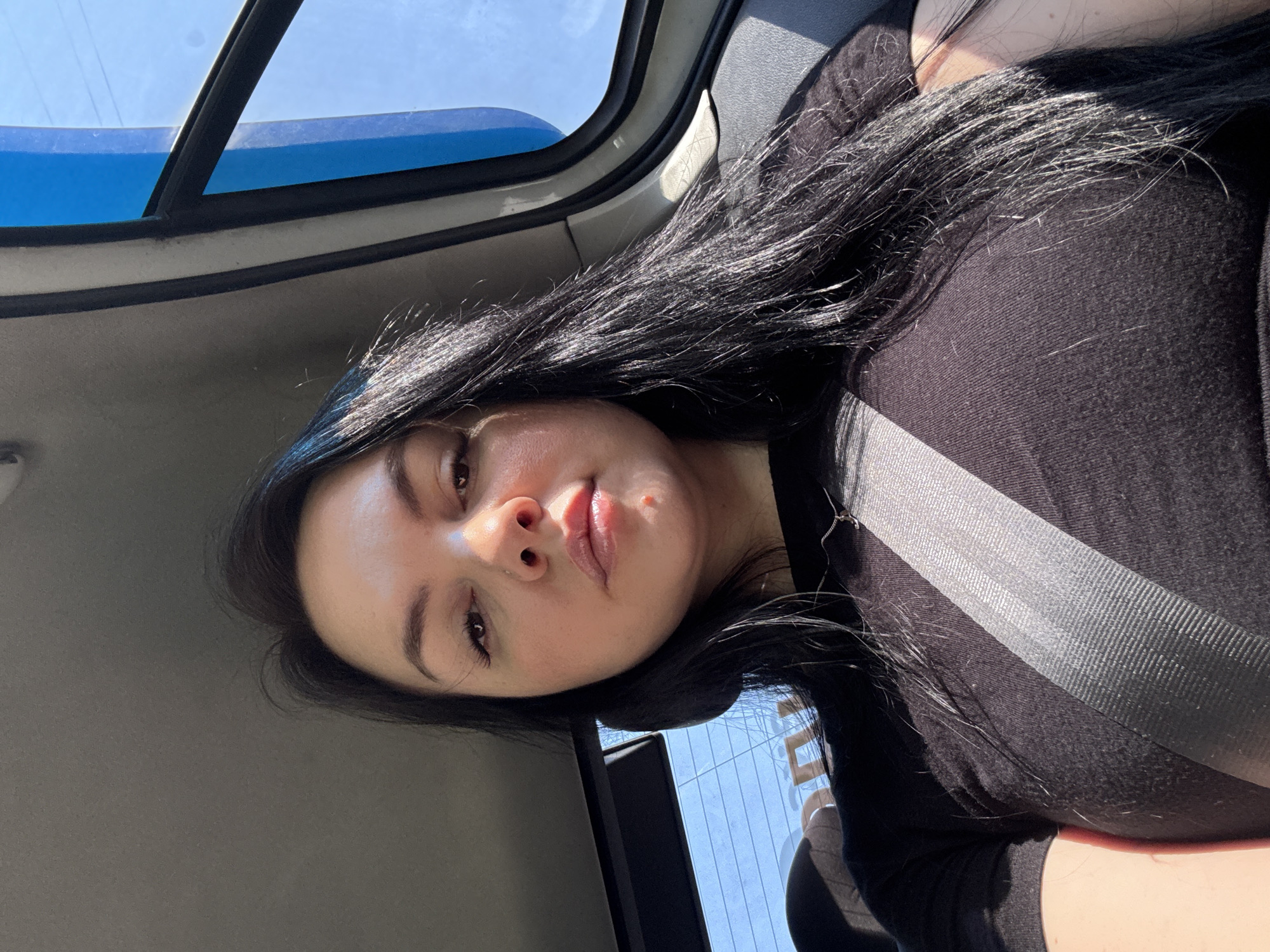 The image shows a woman inside a car, wearing a black top and a seatbelt. The car has a sunroof, through which a glimpse of the sky is visible. The interior of the car is gray. The woman has long black hair, light skin, and appears to be looking at the camera. Given the constraints of the image, it is not possible to pinpoint the exact location in Mytishchi, Russia.