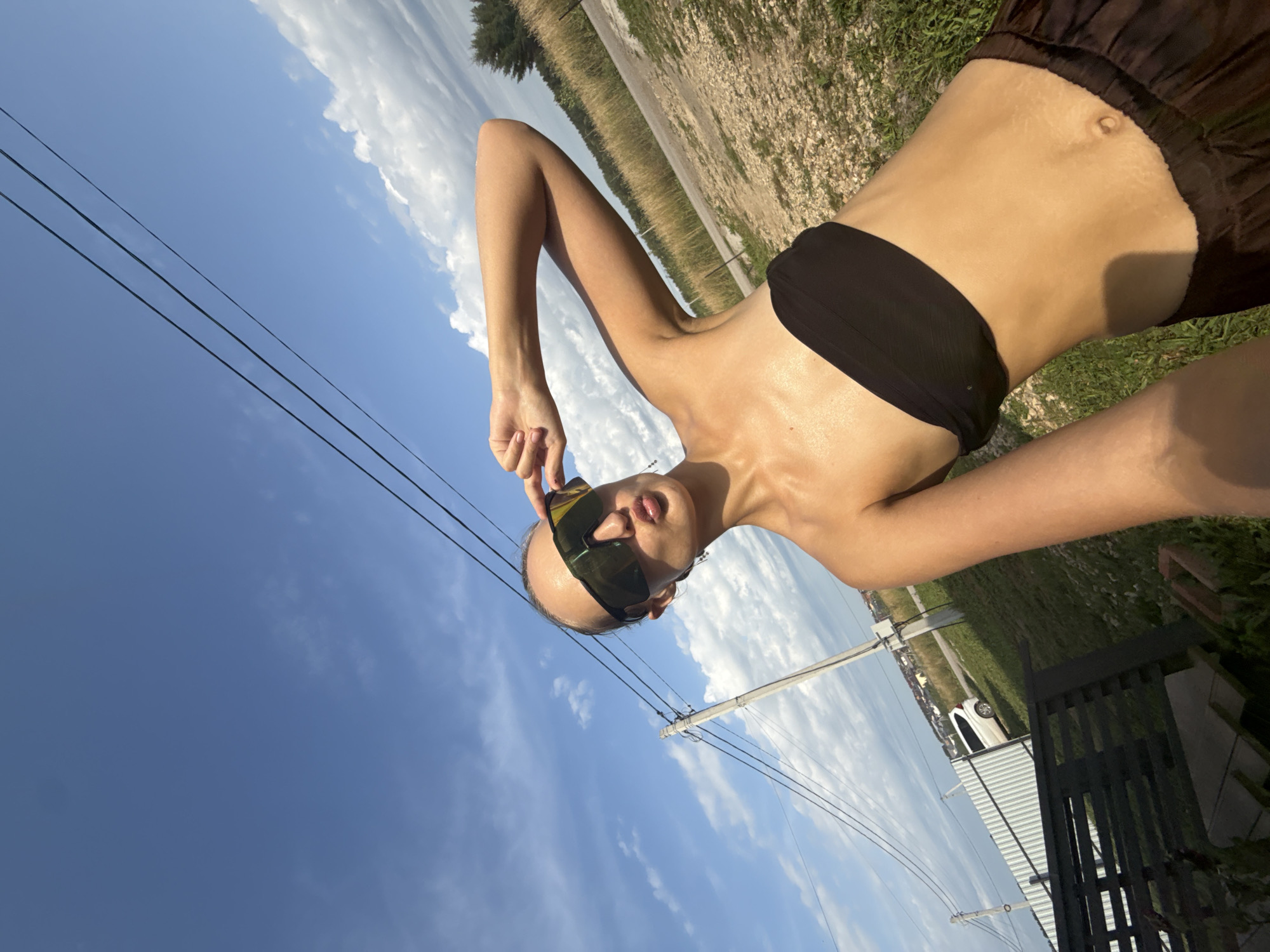 A young woman with her hair pulled back and wearing sunglasses and a black bandeau top stands outdoors in a grassy area with a dirt road in the background. She is holding her right hand up to the side of her face and has her lips puckered. A white car is parked behind a metal fence to her left. Utility poles and wires are visible in the sky. The location is identified as Podshivalovo, Russia.
