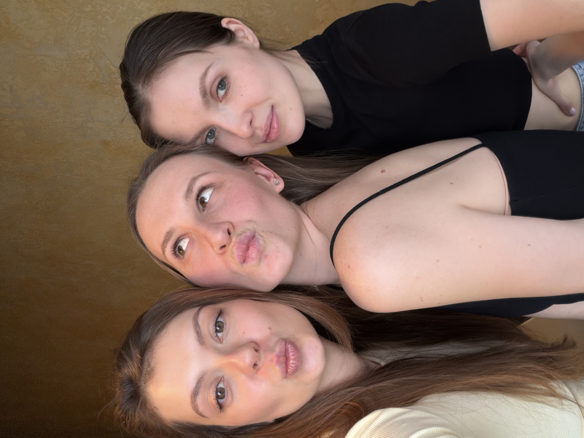 Three women are vertically stacked for a selfie. The top woman has short brown hair and is wearing a black t-shirt. The middle woman has long brown hair and is wearing a black tank top. The bottom woman has long brown hair and is wearing a white sweater. All three women are making kissing faces. The background is a blurred yellow-tan color. No evidence suggests this photo was taken in Khimki, Russia.