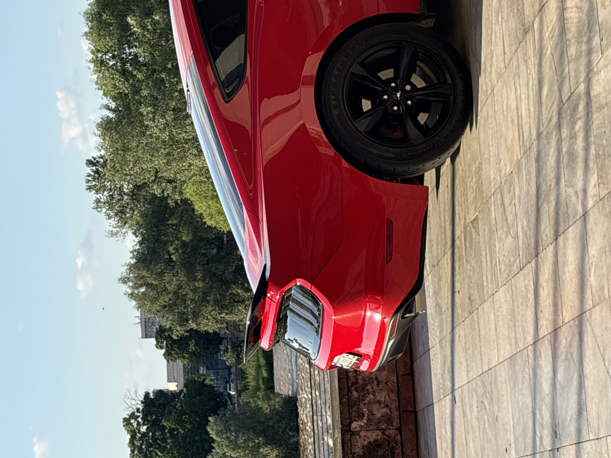 The image shows a red car parked on a tiled surface, likely a sidewalk or public space. The car appears to be a modern muscle car, possibly a Ford Mustang, judging by the taillights and rear quarter panel. The tire is black with a visible logo. To the left of the car, trees suggest a park or green area. Further in the background, one can see buildings, indicating an urban setting. This scene is located in the city of Plovdiv, Bulgaria.