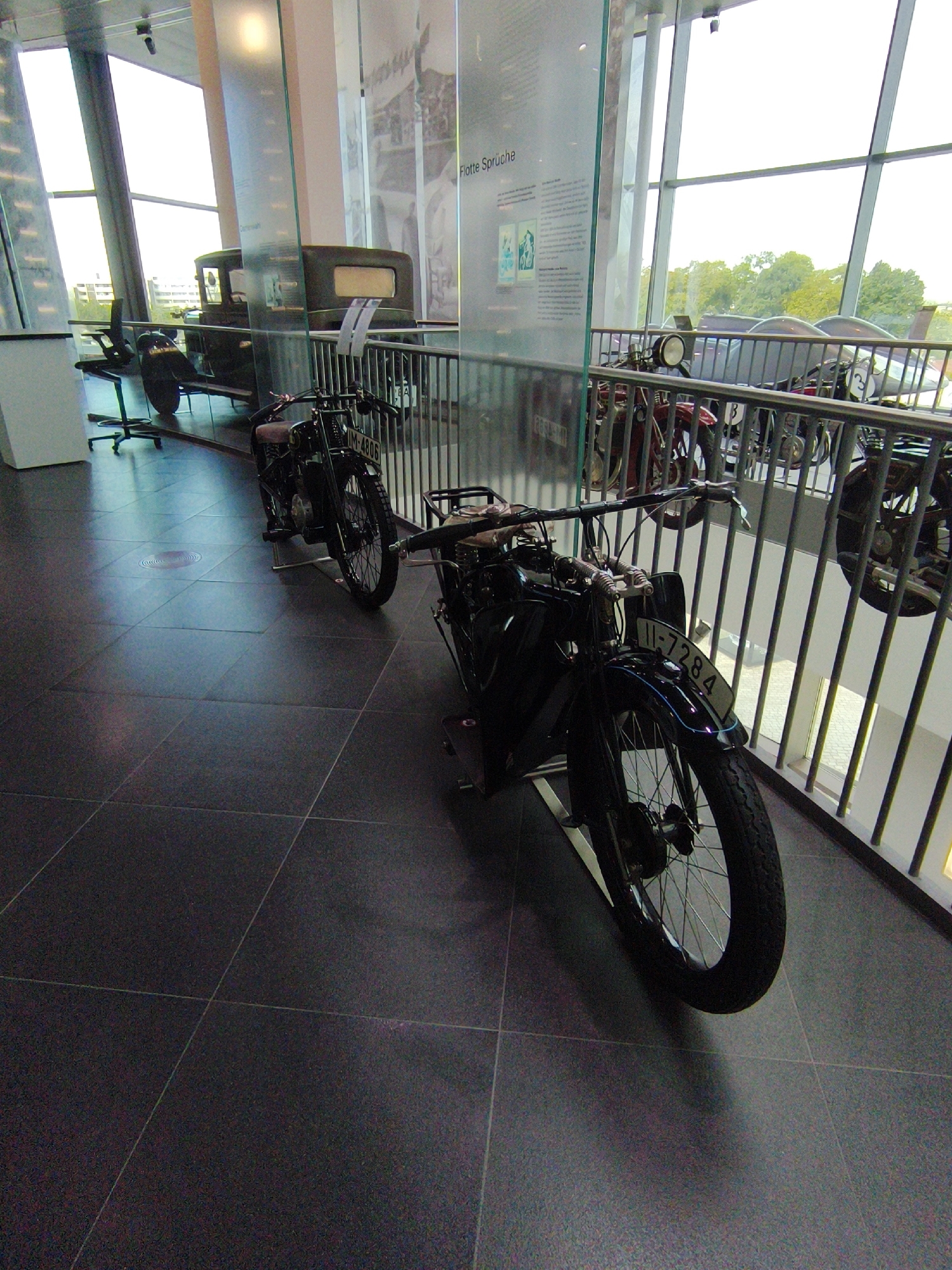 This is a medium-angle shot of a museum exhibit featuring vintage motorcycles and a car, likely taken at the Audi Museum in Ingolstadt, Germany. The motorcycles are parked in a row, with the closest one in sharp focus in the foreground. It is a black motorcycle with a license plate that reads "11-7284." Behind it, another motorcycle is partially visible, and further back, a dark-colored car is parked. The exhibit is set against a backdrop of large windows that reveal trees and buildings outside. A metal railing separates the exhibit from the viewer's area. The floor is tiled in a dark gray.