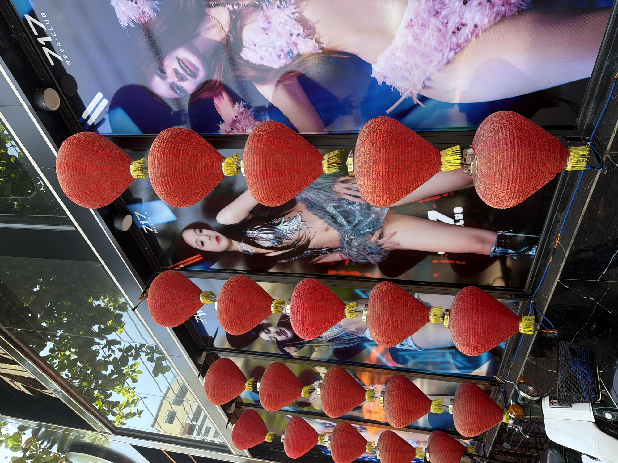 In Nha Trang Ward, Vietnam, a storefront features a display of red Chinese lanterns strung vertically in front of large advertisements. The advertisements depict women in provocative attire, with one clearly showing "717 BEER CLUB" branding. The lanterns, with yellow tassels, hang in multiple rows, creating a festive yet suggestive atmosphere. A white and green scooter is parked in the foreground, partially obscuring the bottom of the display. The building's facade is modern, with large glass windows reflecting the sky and surrounding foliage. The scene appears to be outdoors during daylight.