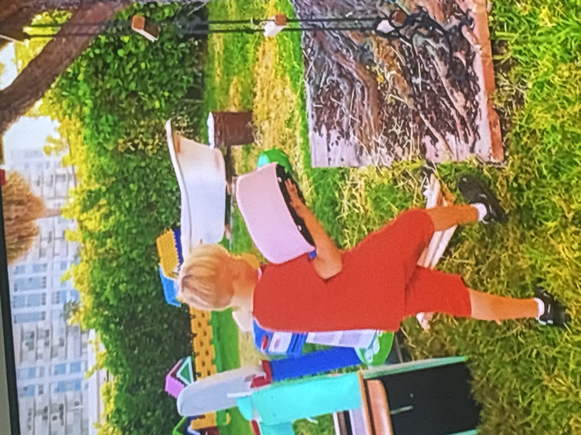 A young boy with blonde hair wearing a red t-shirt and red shorts is holding a pink, round object. He is standing in a grassy outdoor area with a white bathtub and various children's toys visible in the background. The environment appears to be a backyard or play area. The background shows lush green bushes and trees, with buildings visible in the distance. The lighting suggests it is daytime.