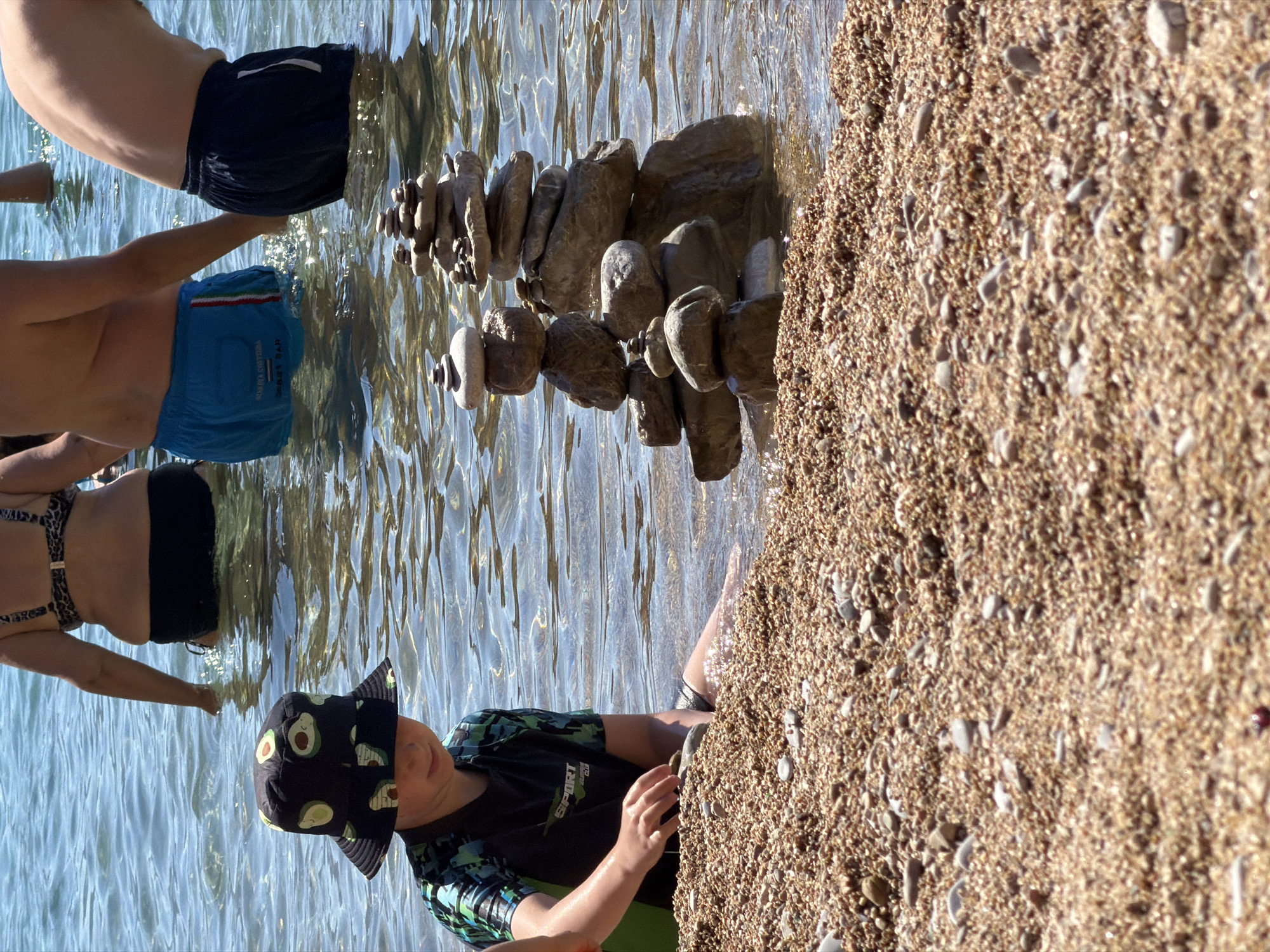 The image shows a group of people at a beach in Budva, Montenegro. The sun is shining, indicating it is daytime and likely warm weather. The water is clear and calm, suggesting a pleasant beach day.
In the foreground, a young boy wearing a black hat with an avocado pattern and a black and green rash guard is sitting on the pebbly shore. He is reaching out with his right hand towards a carefully constructed cairn of stones. The cairn is made of various sizes and shapes of smooth, water-worn rocks, stacked in a precarious balance.
Further back in the water, two adults are partially visible. One is wearing blue swim trunks, and the other is wearing a black bikini. They appear to be standing or wading in the shallow water. The water reflects the sunlight, creating a shimmering effect.
The background consists of the clear water and the pebbly shore, which is densely packed with small stones of various colors. The overall scene depicts a relaxing day at the beach, with the boy engaged in the creative activity of building a stone cairn. The image conveys a sense of summer enjoyment and playful interaction with nature. There is some visible text on the boy's rash guard, but it is partially obscured and difficult to read clearly, appearing to be "SPORT" and possibly other words.