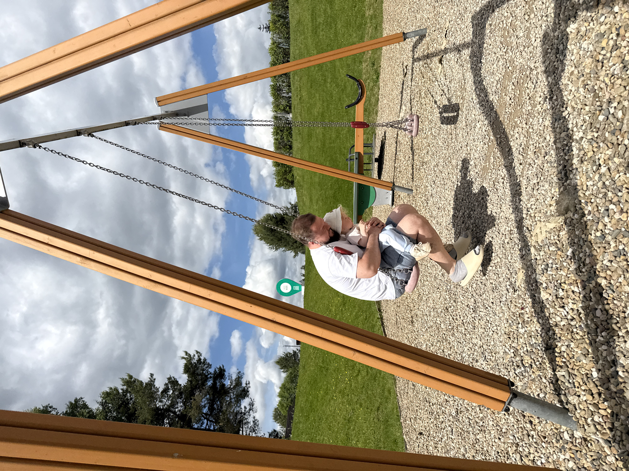 The image captures an intimate moment at an outdoor playground, likely in Dobrograd, Russia, on a bright, partly cloudy day. A man, dressed in a white t-shirt and jeans, is seated on a swing, gently holding a young child, possibly an infant or toddler, who is wearing a light-colored hat. The child is nestled securely in the man's lap.
The scene is set within a playground area featuring a modern swing set constructed from sturdy wooden beams and metal chains. The ground beneath the swings is covered with light-colored gravel, where prominent shadows are cast, indicating clear daylight. Beyond the playground area, expansive green grass and a line of trees are visible under a sky dotted with fluffy white clouds. In the background, another empty swing with both a U-shaped seat and a pink bucket seat can be seen. A circular green sign with white text or an icon, possibly "50" and other symbols, is visible on the distant grass. The image conveys a sense of calm and parental care within an open, natural environment.