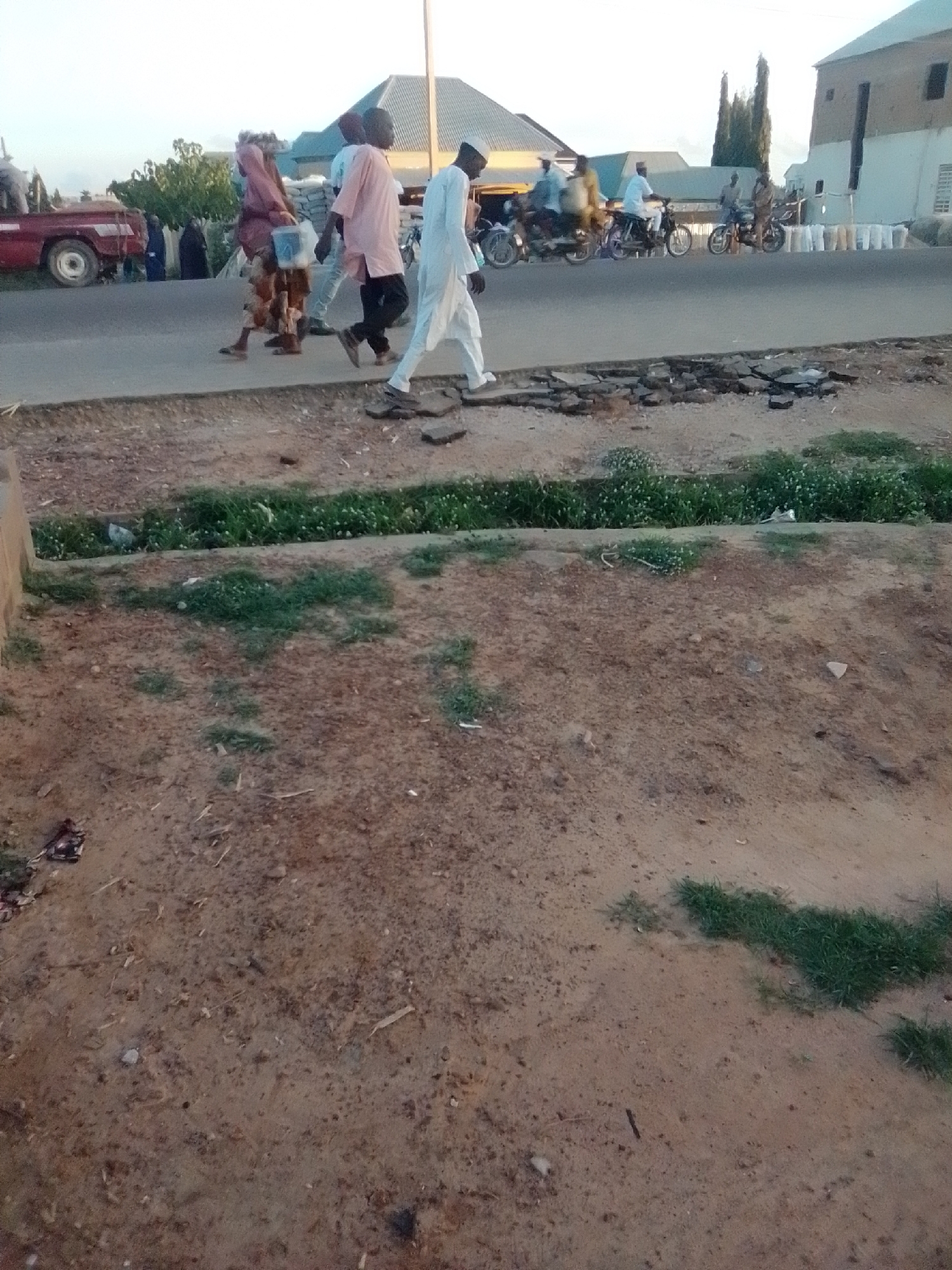 The image captures a street scene in Gombe, Nigeria. In the foreground, a dirt path is visible with scattered debris and patches of grass. A small drainage channel with lush green vegetation runs along the edge of the path.
In the midground, a paved road is present. Several individuals are walking along the side of the road. One man in a white traditional outfit and cap is walking towards the right of the frame. He is followed by a group of people, including one in a light pink shirt and dark pants, and another in a patterned garment with a bucket. Further down the road, several motorcycles with riders are visible, some in motion, suggesting ongoing traffic. The background features buildings with distinct architectural styles, including a prominent structure with a light-colored facade and a darker, possibly metal, roof. The sky above is bright, indicating daylight. The overall impression is a lively street with pedestrian and vehicular activity.