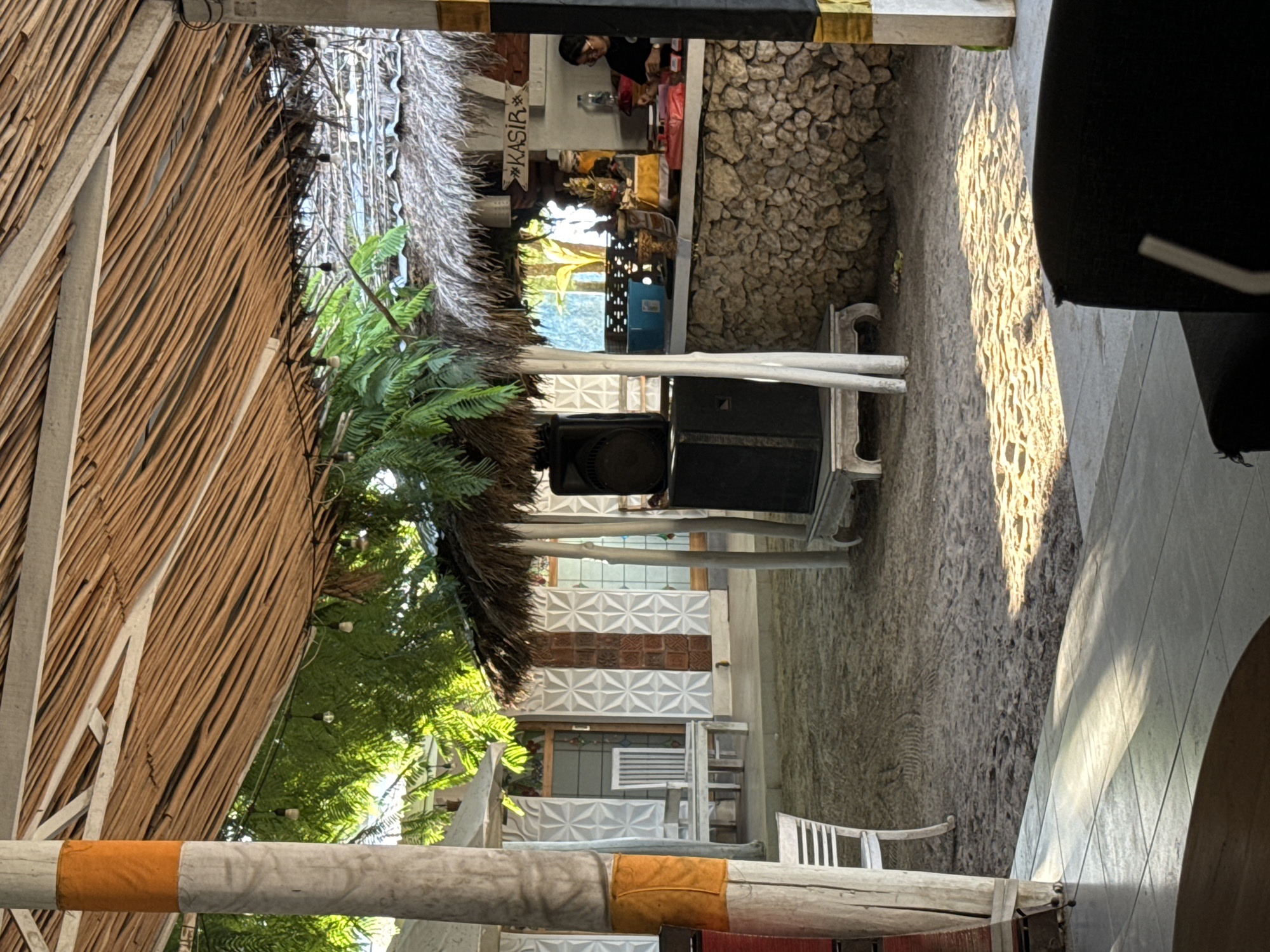 The image depicts an inviting outdoor or semi-outdoor restaurant or cafe setting in Nusa Penida, Indonesia. The venue features a rustic, tropical design with a prominent thatched roof and walls constructed from woven bamboo or thin wooden sticks, supported by white pillars.
In the mid-ground, a cashier station is visible, marked by a hanging sign that clearly reads "KASIR" (Indonesian for Cashier). Behind this counter, a staff member is partially seen, attending to items on the counter, which include traditional offerings (yellow/orange cloth items) and other supplies. Lush green ferns and tropical plants are abundant, adding to the natural and serene atmosphere. Overhead, string lights are visible, suggesting the venue is used from day into evening. Two large black speakers are stacked on a white stand, indicating potential for music or events.
The ground surface is a mix of natural rock and paved areas, with striking patches of bright sunlight and strong shadows, characteristic of either early morning or late afternoon light. The warm, dappled sunlight creates a pleasant glow across the scene. White chairs and tables are arranged throughout the space, including a distinct white bench in the foreground on the left. A decorative wall with white and brown patterned tiles adds an artistic touch to the architecture. The overall impression is one of a relaxed, tropical leisure spot.